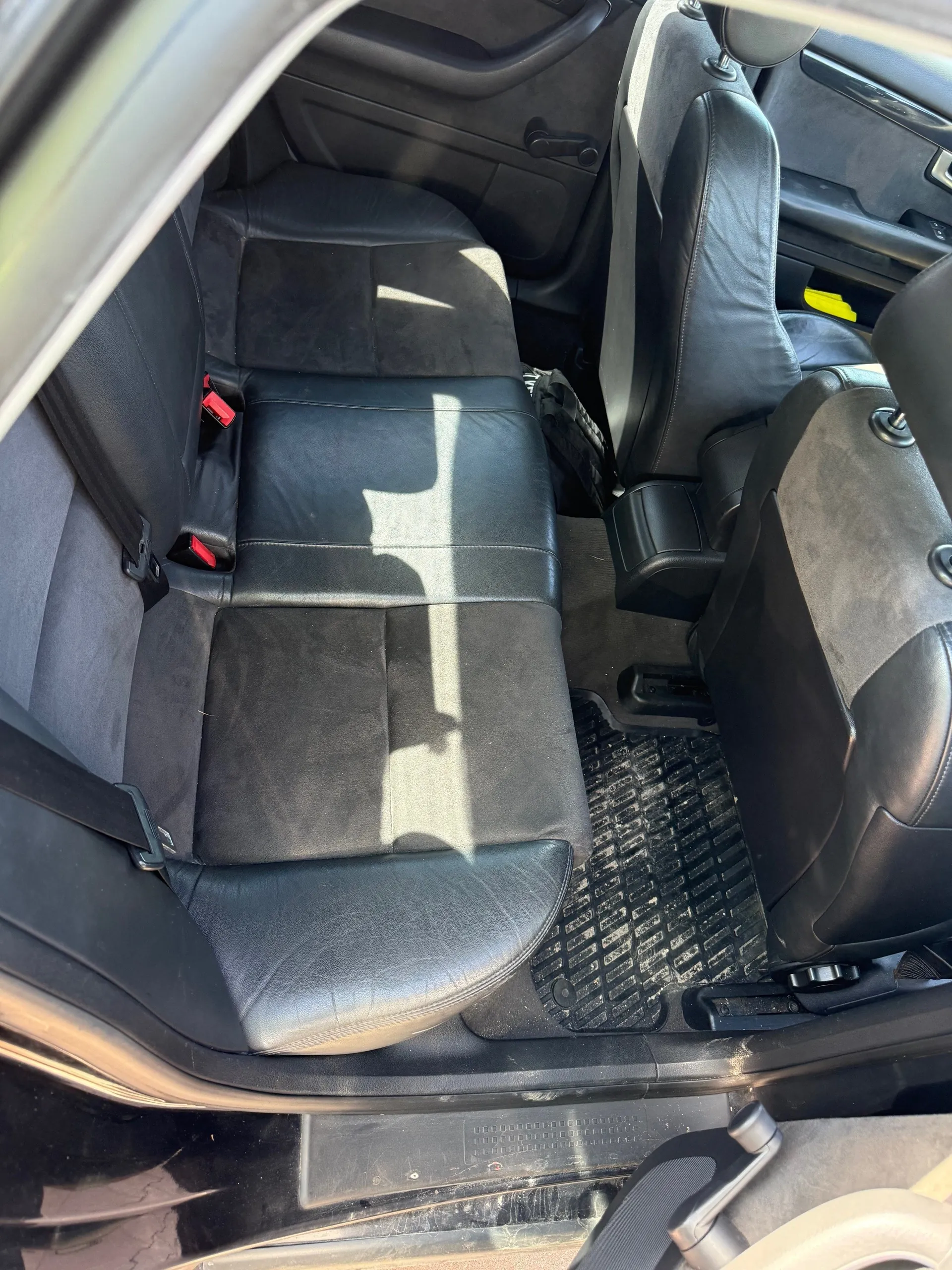 Wnętrze samochodu z widokiem na czarne siedzenia tylnego rzędu z czarną skórzaną i sukienną tapicerką. Widoczne są czerwone klamry pasów bezpieczeństwa, czarna gumowa mata na podłodze oraz fragmenty przednich siedzeń. W tle widać otwarte drzwi samochodu i fragment deski rozdzielczej z żółtym elementem. Na siedzeniach pada cień od okna.