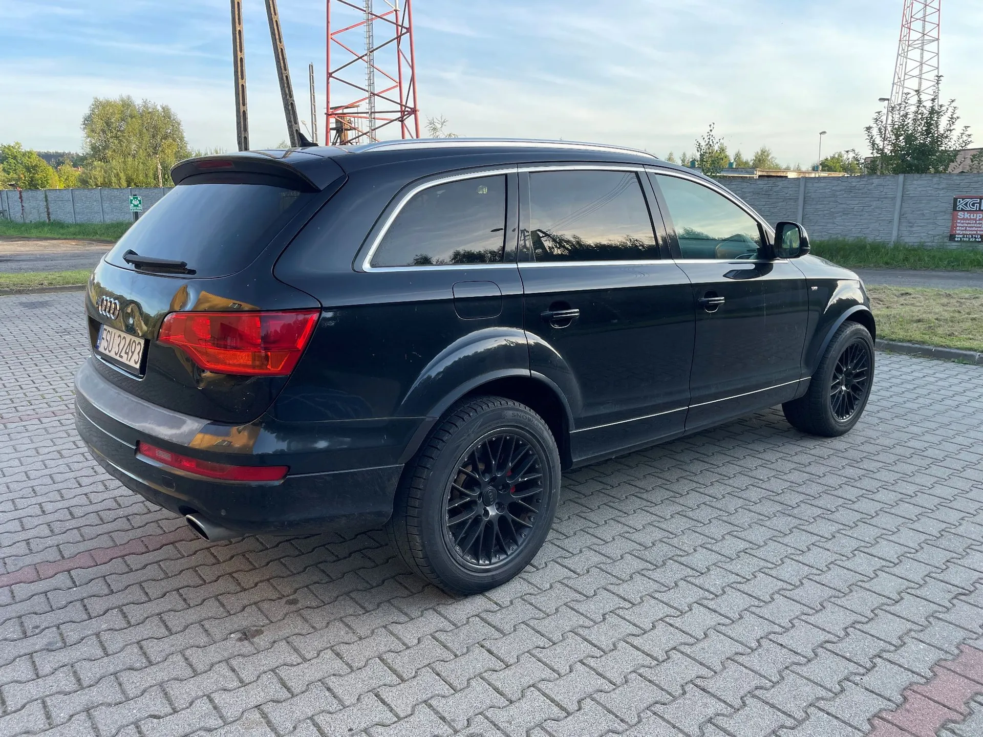 Czarny samochód marki Audi typu SUV zaparkowany na kostce brukowej. Widok z tyłu i z boku. Pojazd ma czarne felgi i tablicę rejestracyjną o numerze SU 32493. W tle widoczna jest wysoka, czerwono-biała konstrukcja masztu telekomunikacyjnego oraz betonowy płot.