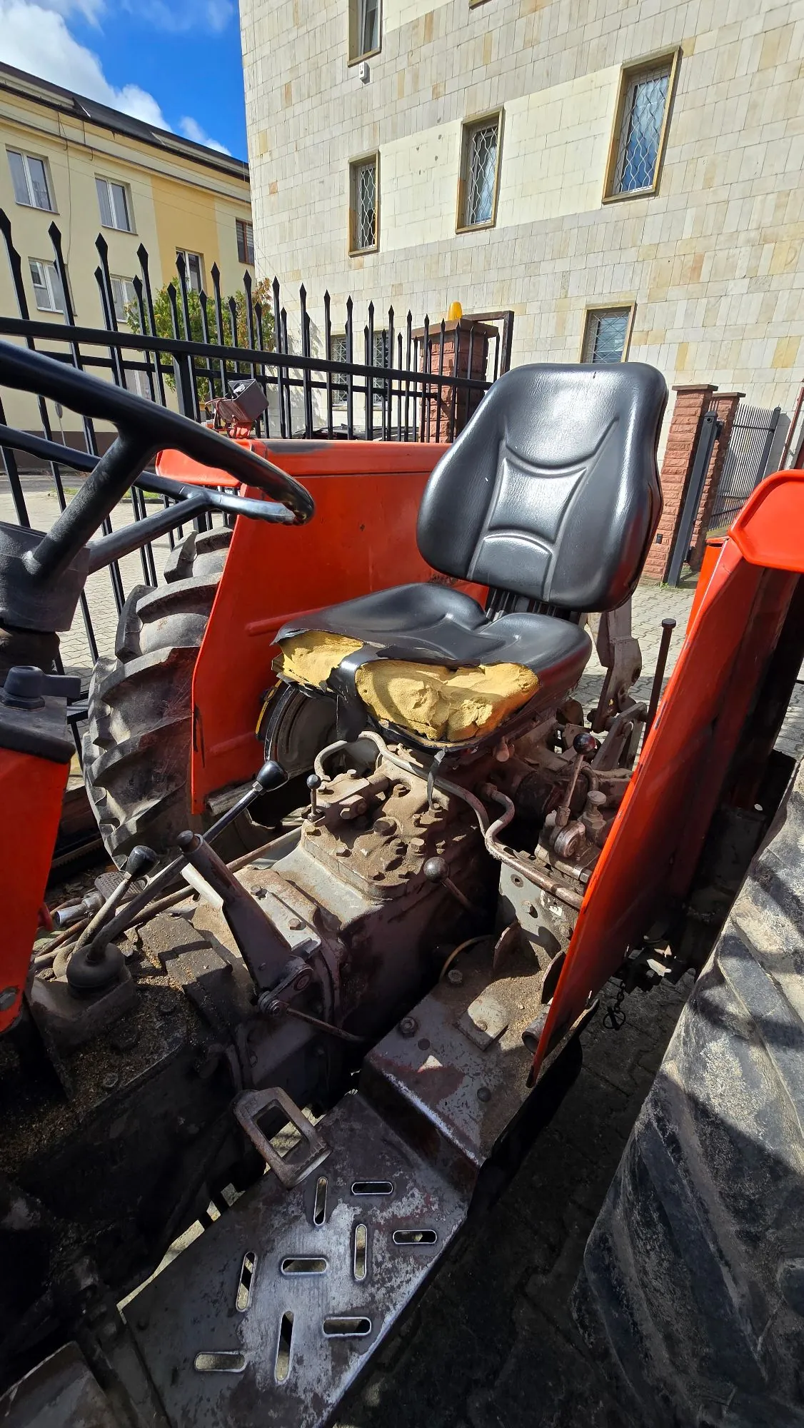 Zbliżenie na czerwony traktor z czarnym, plastikowym siedzeniem, którego dolna część tapicerki jest przerwana, odsłaniając żółtą gąbkę. Widoczna jest metalowa konstrukcja maszyny, dźwignie oraz fragmenty dużych, błotnistych opon. W tle znajduje się czarny metalowy płot oraz jasny budynek z oknami.
