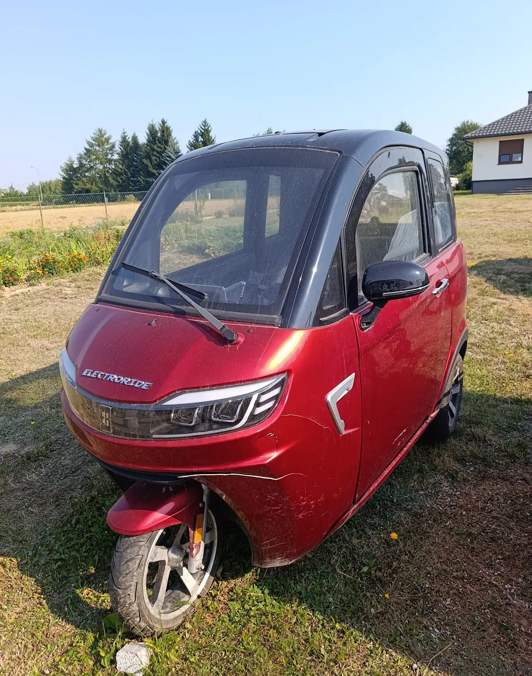 Czerwony, niewielki samochód elektryczny marki Electroride zaparkowany na trawiastym terenie. Pojazd ma czarny dach i czarne lusterka boczne. Na przodzie widnieje napis 'ELECTRORIDE'. W tle widać zielone drzewa, płot oraz fragment białego budynku z brązowym dachem pod jasnym, bezchmurnym niebem.