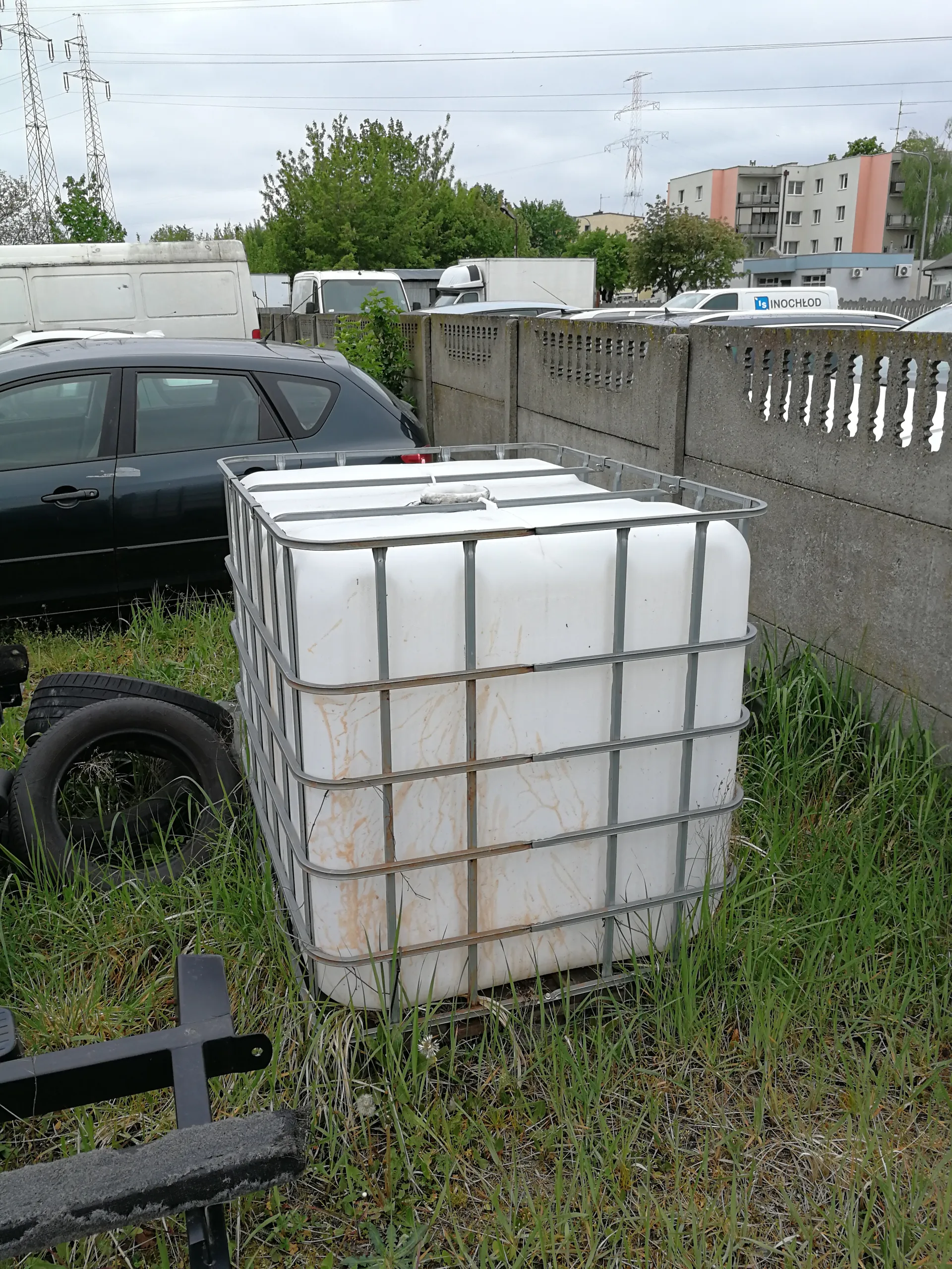 Biały zbiornik IBC w metalowej ramie z widocznymi plamami rdzy, umieszczony na porośniętym trawą terenie. Obok zbiornika leżą dwa stare opony. Za zbiornikiem zaparkowany jest czarny samochód osobowy. W tle betonowy płot z ozdobnymi wycięciami, za którym widoczne są pojazdy, w tym biały furgon z napisem INOCHLOR. Dalszy plan: budynki mieszkalne, drzewa, linie energetyczne i maszty pod chmurnym niebem.