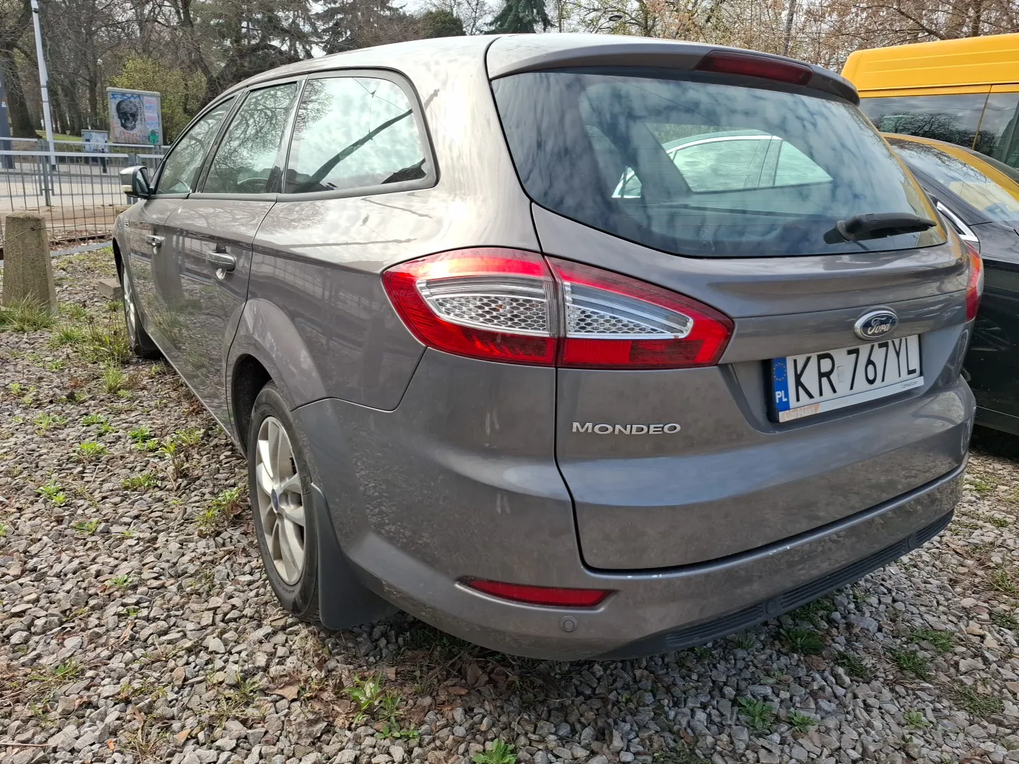 Szary samochód Ford Mondeo zaparkowany na żwirowym placu. Widoczny tył pojazdu z tablicą rejestracyjną KR 767YL (PL) oraz napisem "MONDEO" na bagażniku. Obok znajduje się czarny samochód i żółty pojazd. Tło: drzewa, ogrodzenie i tablica informacyjna.