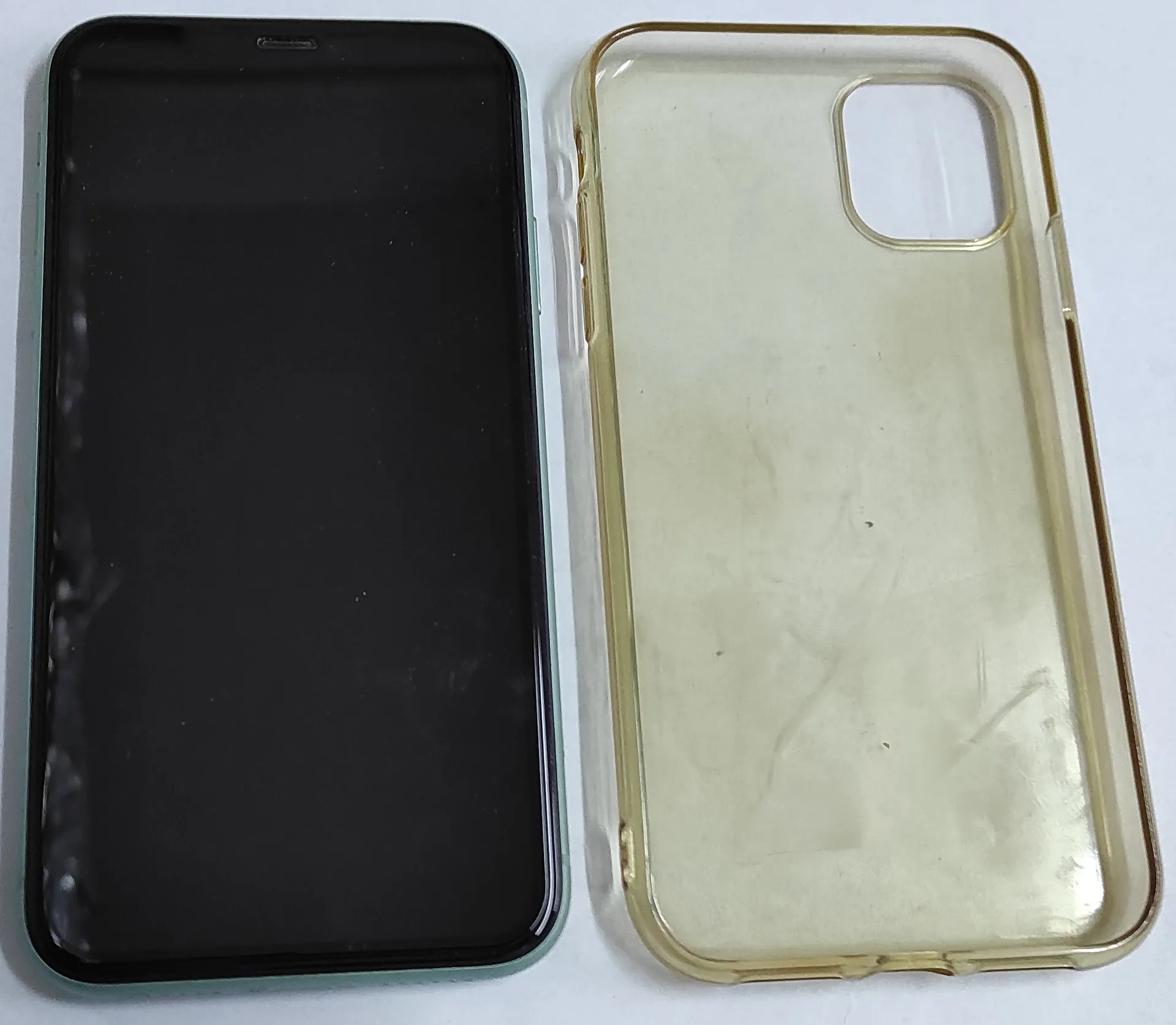 Obok siebie leżą dwa przedmioty: po lewej stronie czarny smartfon z wyłączonym ekranem, a po prawej przezroczyste, lekko zżółknięte silikonowe etui ochronne z wycięciem na aparat w prawym górnym rogu.