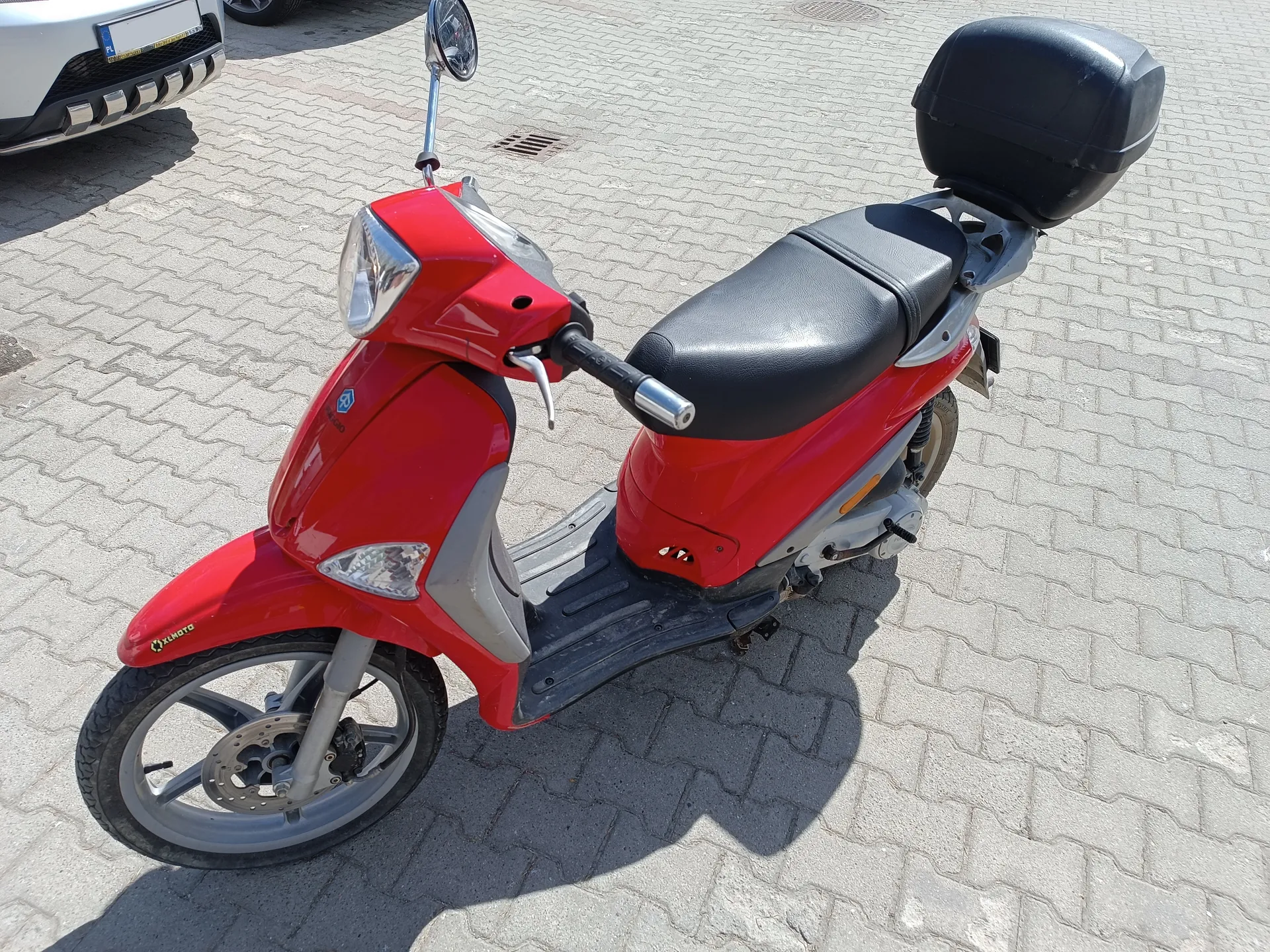 Czerwony skuter z czarnym siedzeniem i czarnym bagażnikiem na tylnej części, zaparkowany na brukowanej powierzchni. Skuter posiada przednią i tylną oświetlenie, lusterko wsteczne oraz widoczny napis 'X-MOTO' na przedniej części. W tle widoczny jest fragment białego samochodu z tablicą rejestracyjną oraz kanałowy żłobek na chodniku.