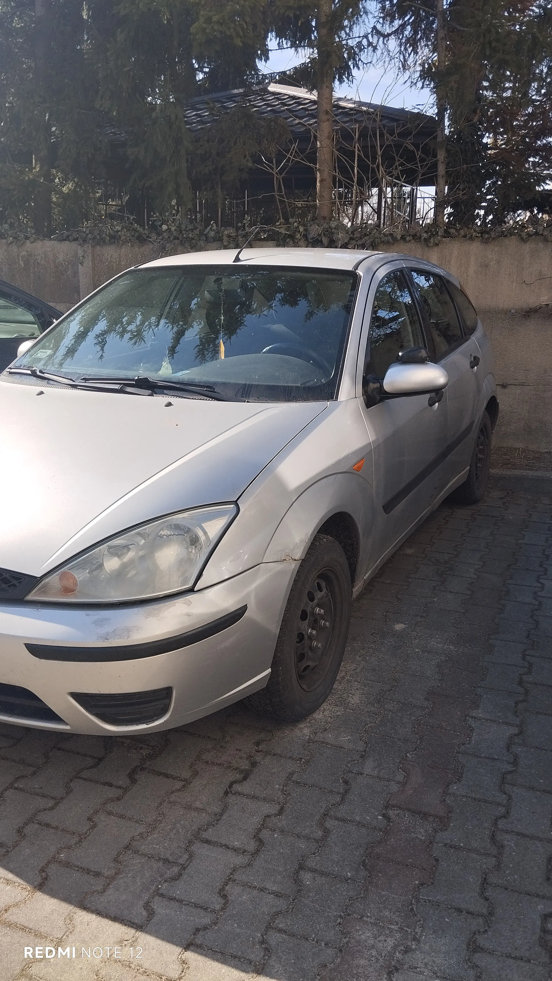 Srebrny samochód osobowy typu hatchback zaparkowany na brukowanej powierzchni. W tle betonowy mur z zielonymi pnączami, drzewa oraz budynek z ciemnym dachem. Po lewej stronie częściowo widoczny inny samochód. W dolnym lewym rogu obrazu widoczny napis "REDMI NOTE 12". Samochód ma czarne felgi, przednie światła i lusterko boczne po prawej stronie.