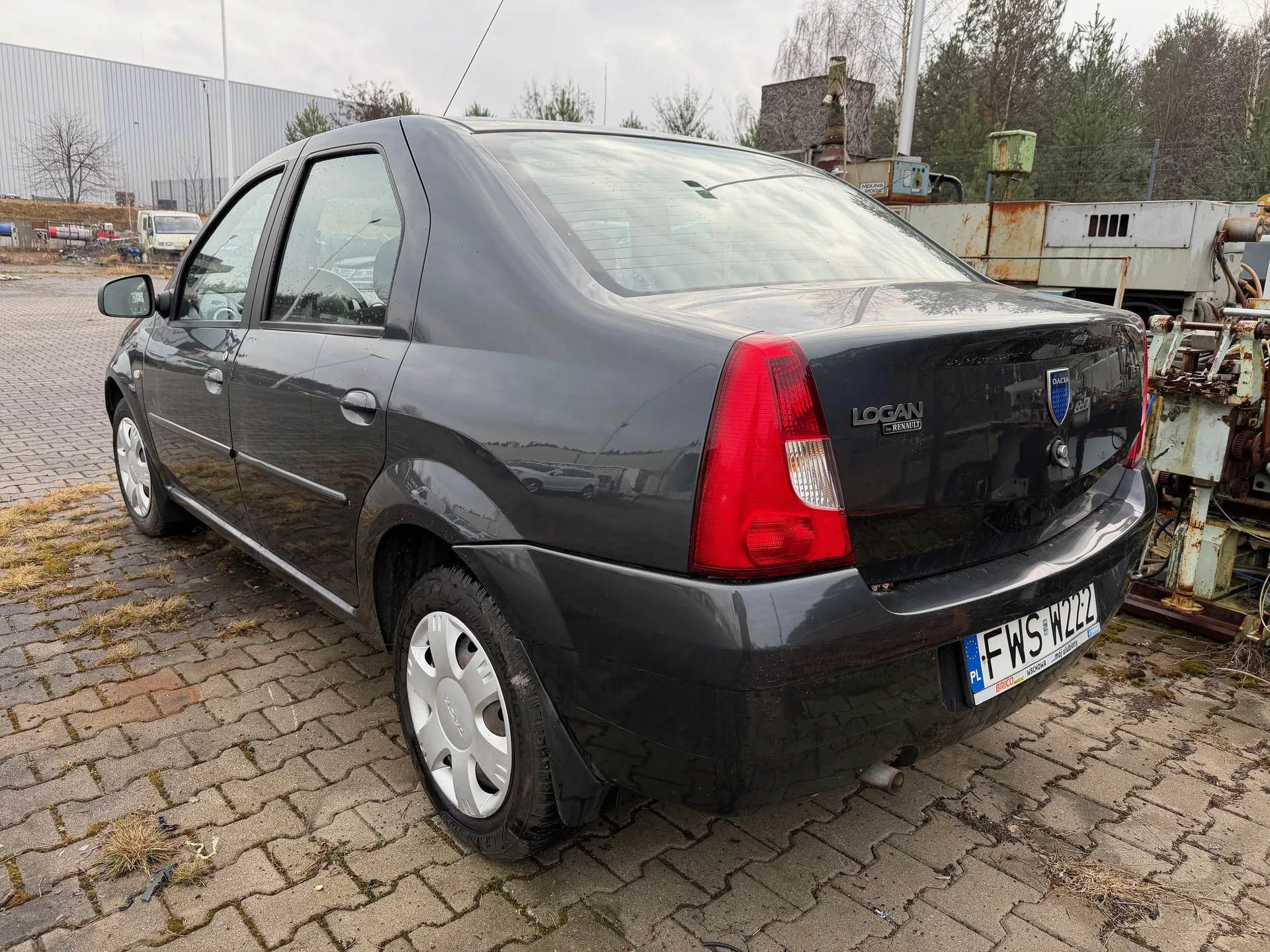 Czarny samochód osobowy Dacia Logan z widocznym napisem "LOGAN" i logo marki na tylnej klapie. Tablica rejestracyjna oznaczona numerem FWS W222 z kodem kraju PL. Samochód zaparkowany na brukowanej powierzchni z fragmentami trawy, na tle przemysłowych maszyn, budynku i drzew. Niebo jest zachmurzone.