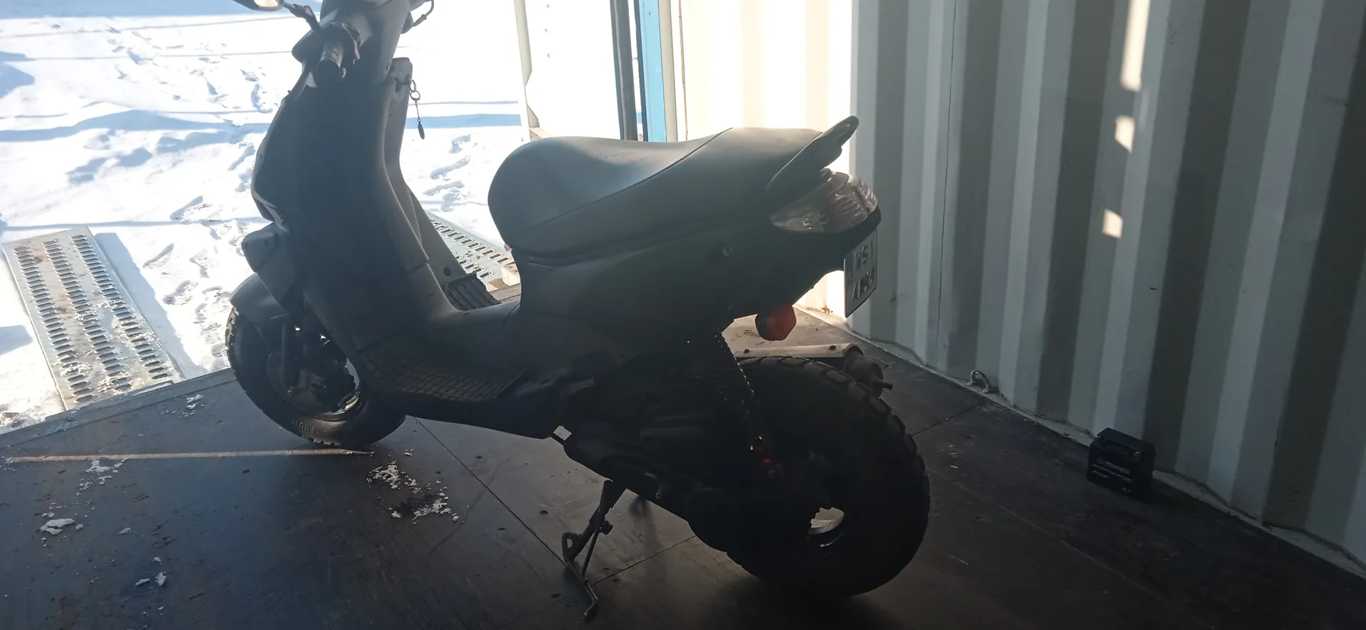 Czarny skuter zaparkowany wewnątrz metalowego kontenera z pionowymi listwami. Skuter ma czarne siedzenie, tablicę rejestracyjną z tyłu oraz podniesioną podpórkę. W tle widoczny jest śnieżny krajobraz z metalowymi kratami na podłodze. Na podłodze obok ściany kontenera znajduje się czarne pudełko z napisem "MOTOBATT". Podłoga kontenera jest ciemna z rozsypanymi resztkami śniegu.