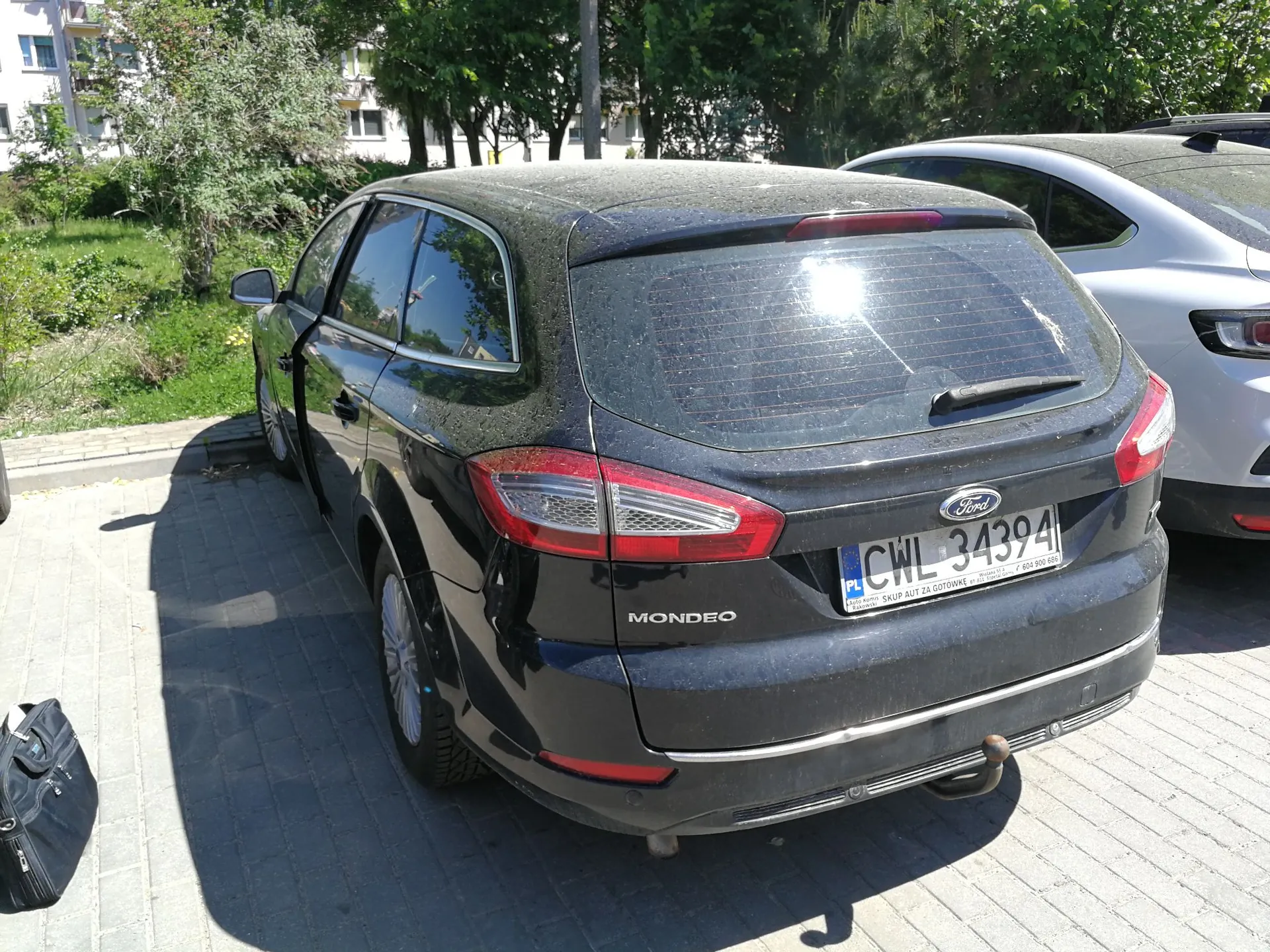 Czarny samochód marki Ford Mondeo w nadwoziu kombi, zaparkowany tyłem na kostce brukowej. Pojazd jest zakurzony. Widoczna jest tablica rejestracyjna o numerze CWL 34394 z oznaczeniem PL. W tle widać fragment innego, srebrnego samochodu oraz zielone drzewa i fragmenty budynków mieszkalnych.