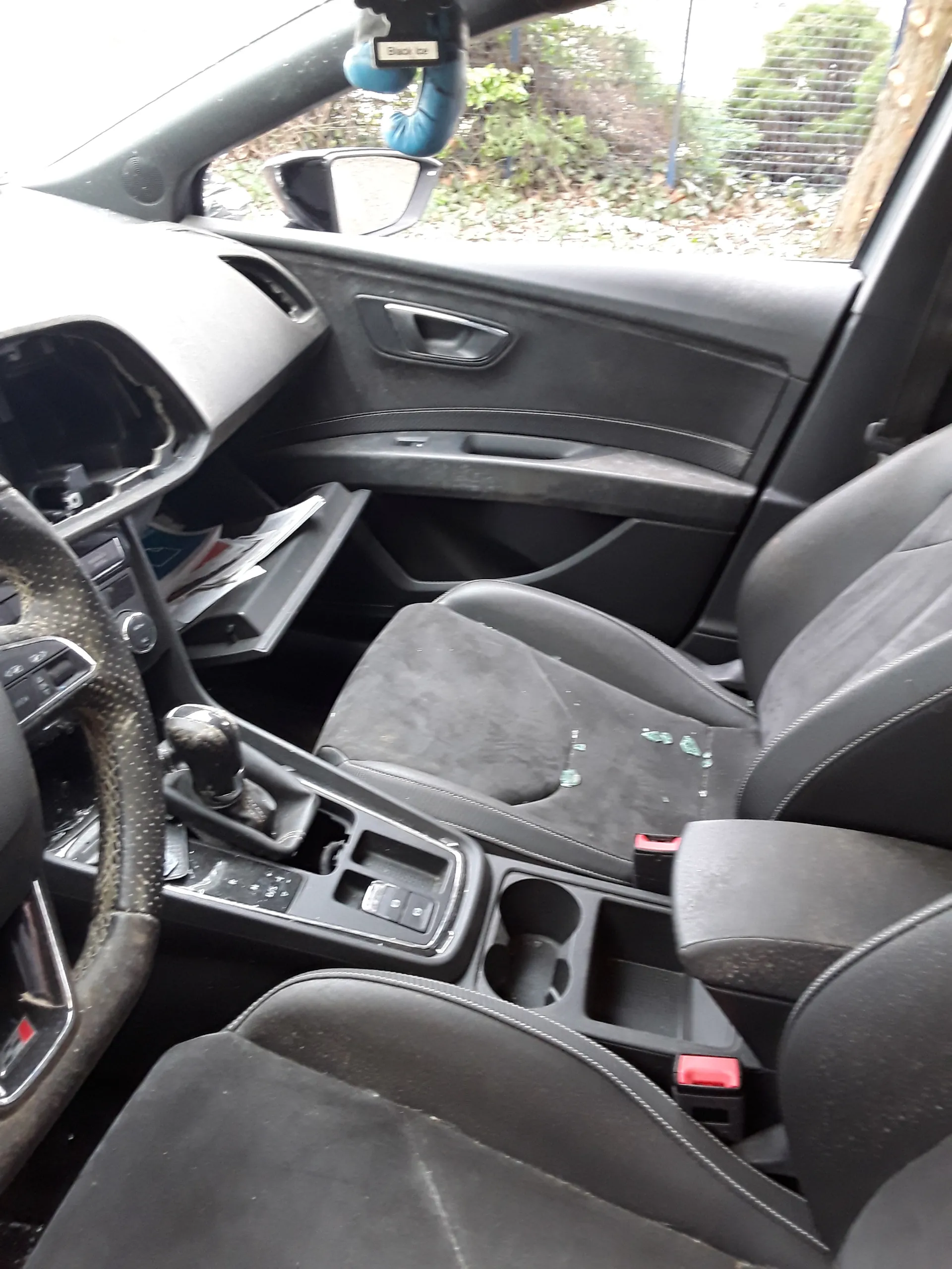 Wnętrze samochodu z czarnymi siedzeniami, centralną konsolą z dźwignią zmiany biegów, uchwytem na kubki i przyciskami. Widoczne są drzwi z uchwytem i oknem, przez które widać siatkę ogrodzeniową oraz zieloną roślinność. Na siedzeniu pasażera znajdują się zielone plamy. W dolnej części obrazu kierownica z przyciskami. Po lewej stronie ręka w niebieskiej rękawicy trzymająca urządzenie. W przedziale na dokumenty widoczne są papiery.