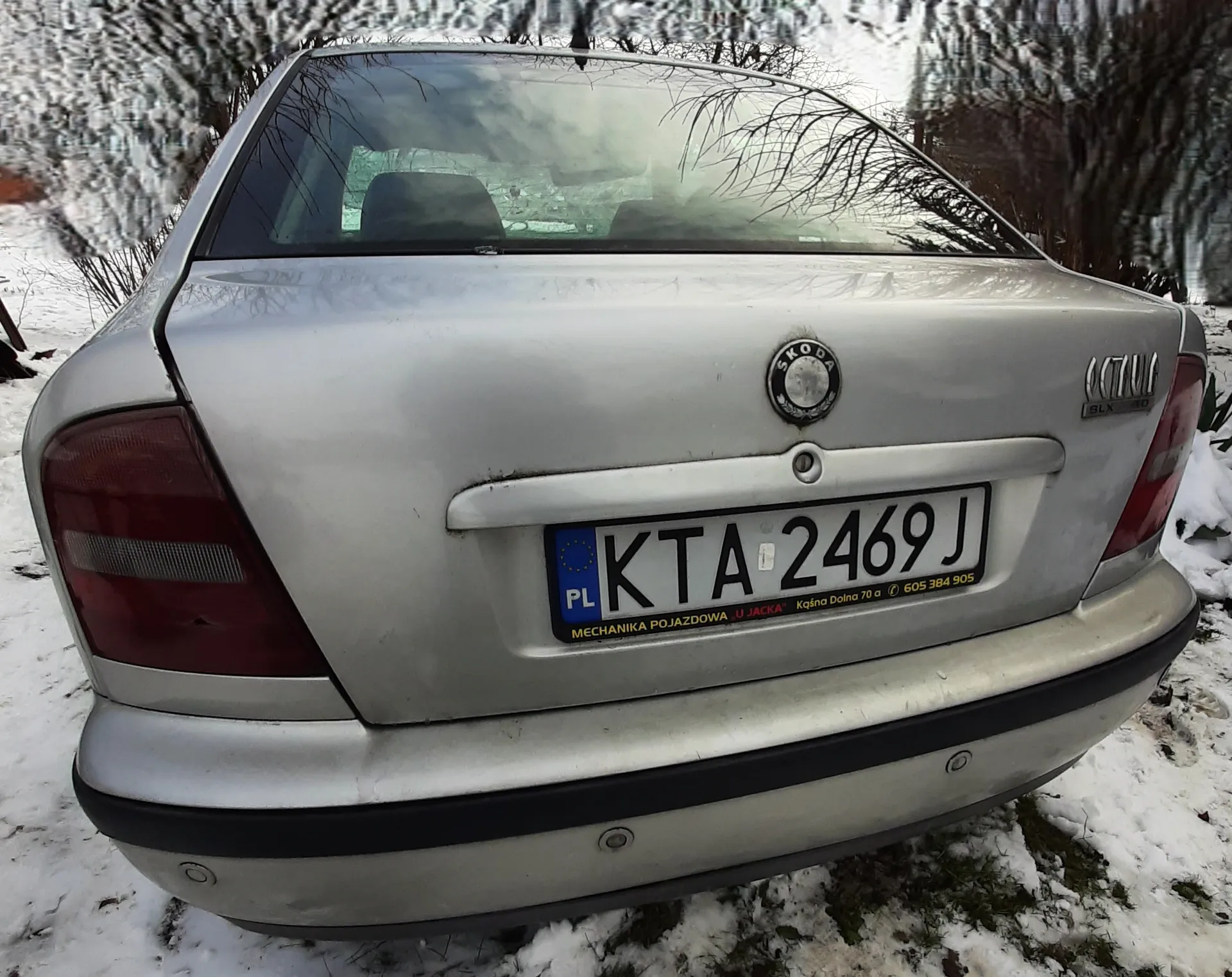 Tył srebrnego samochodu marki Škoda Octavia z tablicą rejestracyjną KTA 2469 J. Na tablicy widoczne są dodatkowe napisy: 'PL', 'MECHANIKA POJAZDOWA U JACKA', 'Kąśna Dolna 70 a' oraz numer telefonu '605 384 905'. Samochód stoi na śnieżnej powierzchni, w tle drzewa bez liści pokryte śniegiem. Na bagażniku widoczny jest emblemat marki i napis 'OCTAVIA'.