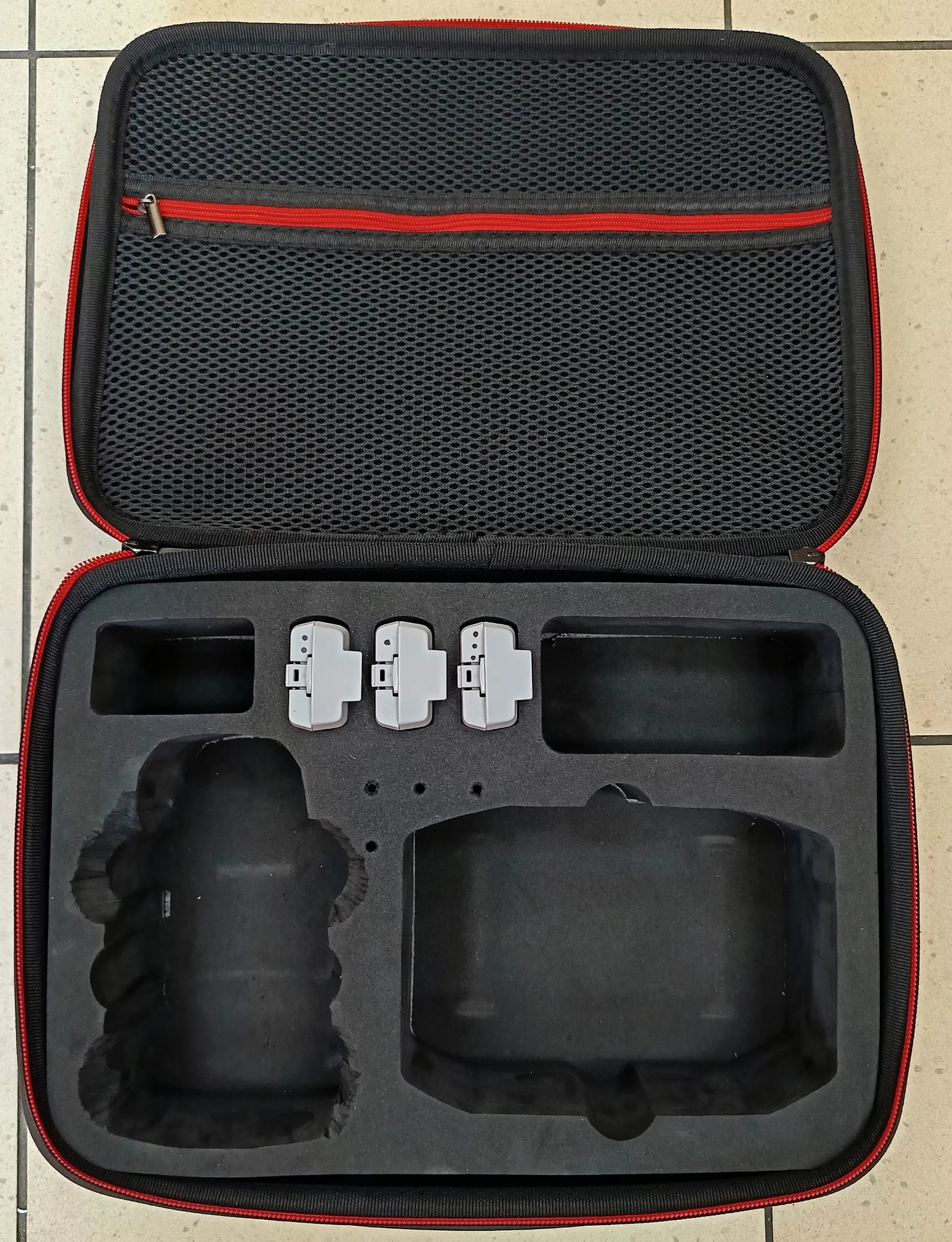Otwarty czarny kufer z czerwonymi elementami, zawierający piankowe przegródki. W środku trzy białe prostokątne elementy z metalowymi detalami, dwa większe czarne przestrzenie na urządzenia oraz siateczkowa kieszonka z czerwonym zamkiem błyskawicznym. Kufer leży na jasnej podłodze z kafelkami.