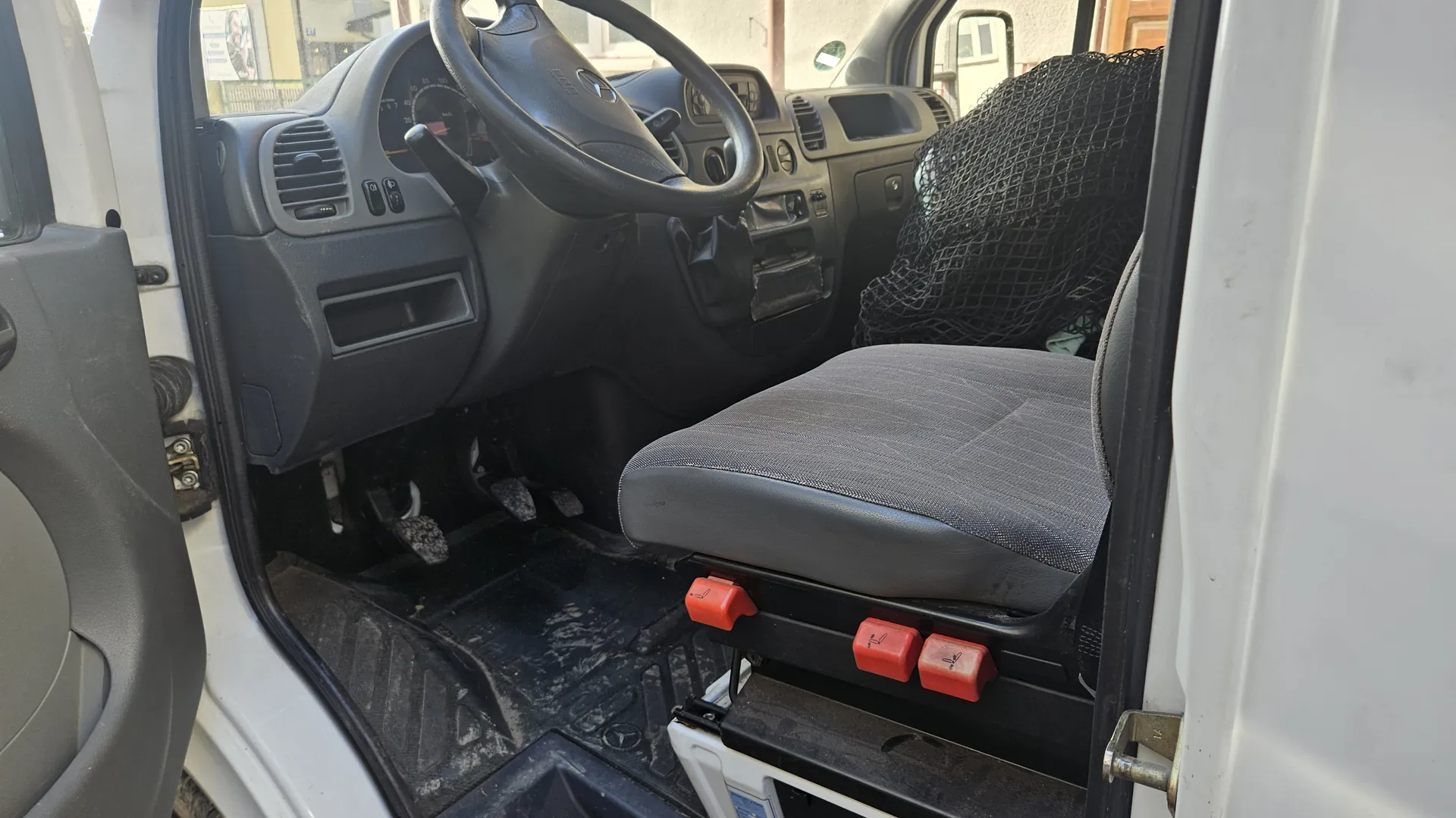 Wnętrze pojazdu widziane z otwartego drzwi kierowcy. Widoczna czarna kierownica z logo, deska rozdzielcza z licznikami, wentylacjami i przyciskami. Siedzenie kierowcy w kolorze szarym z czerwonymi dźwigniami regulacji. Czarne siatki za siedzeniem, brudne dywany podłogowe oraz biały nadwozie pojazdu. W tle przez przednią szybę widoczne budynki i ulica.