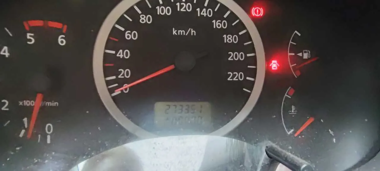 Zbliżenie na deski rozdzielcze samochodu. W centrum znajduje się prędkościomierz z zakresem do 220 km/h, wskazówka na 0 km/h. Poniżej wyświetlacz cyfrowy z przebiegiem 273351. Po lewej stronie tachometr z zakresem do 6 x1000 obr./min, wskazówka na ok. 1. W prawej części widoczne wskaźniki paliwa i temperatury chłodniczego. Aktywne czerwone lampki ostrzegawcze: symbol wykrzyknika w okręgu, paliwa i baterii. Tło deski rozdzielczej jest ciemne, z widocznymi śladami kurzu.