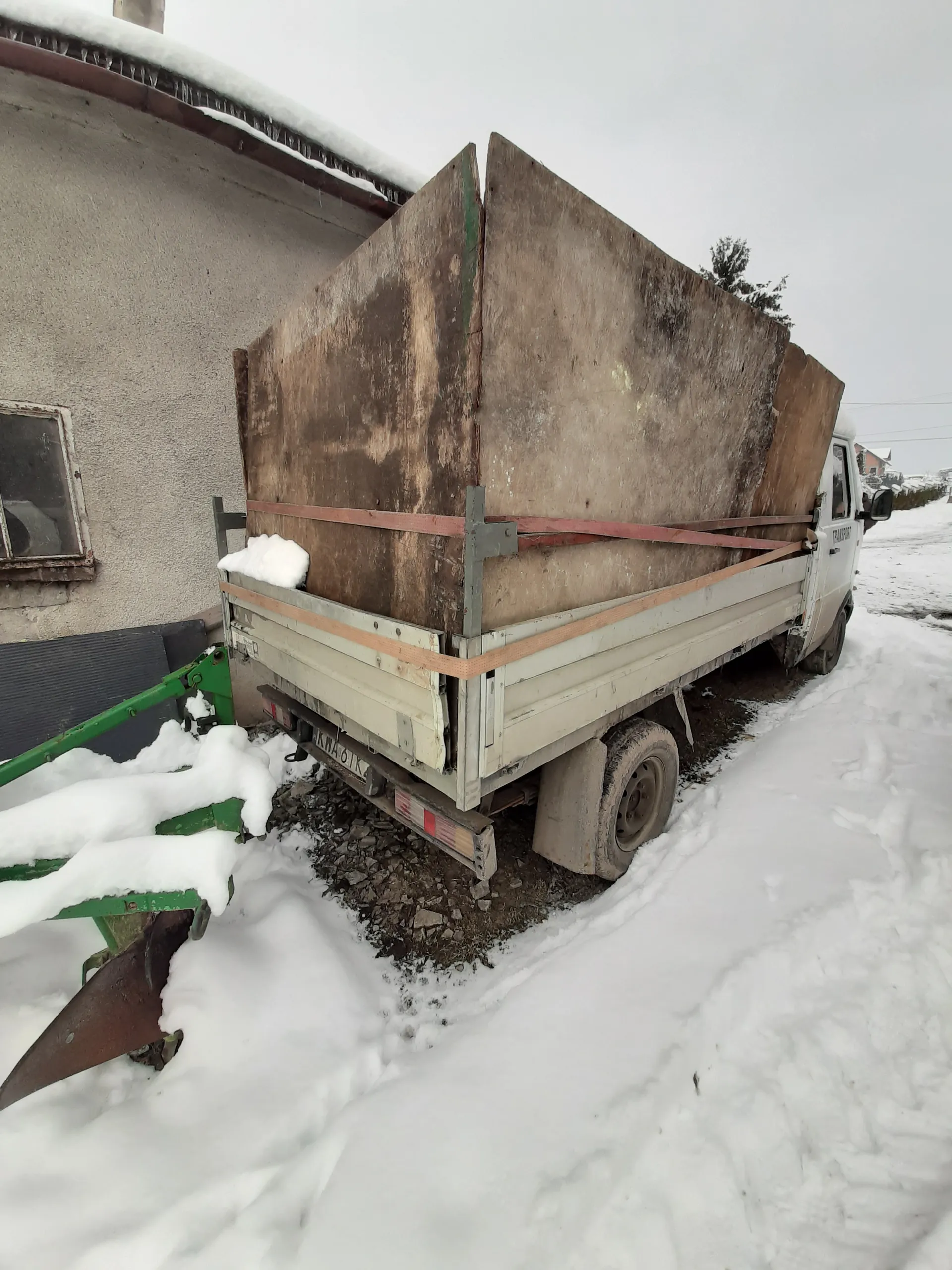 Biały samochód ciężarowy z dużą, zniszczoną drewnianą konstrukcją na platformie, przymocowaną czerwonymi i pomarańczowymi paskami. Po prawej stronie obrazu wysoka zaspy śniegu, po lewej budynek z betonową ścianą i oknem. Na pierwszym planie zielona maszyna pokryta śniegiem. Niebo jest pochmurne, na ziemi widoczne są śnieg i ziemia. Tablica rejestracyjna samochodu nieczytelna.