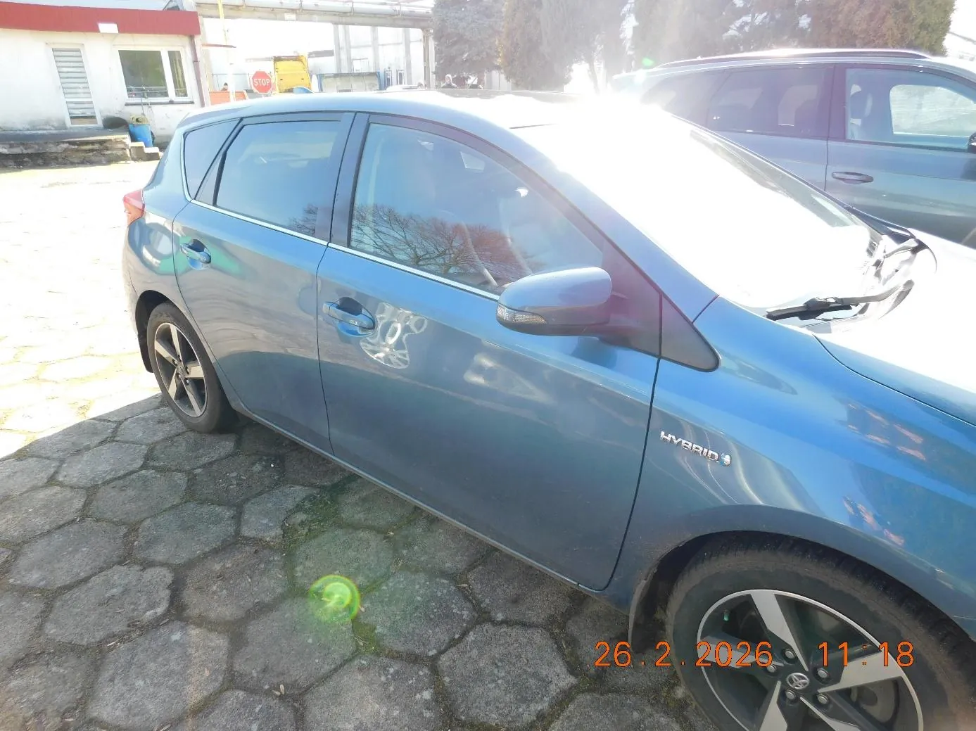 Niebieski samochód hybrydowy zaparkowany na brukowanej powierzchni. Widoczne są boczne drzwi, lusterko boczne oraz napis HYBRID na boku pojazdu. W tle drugi samochód, budynek z czerwonym dachem, drzewa i znak STOP. W prawym dolnym rogu obrazu widoczny pomarańczowy znaczek daty i godziny: 26.2.2026 11:18. Słoneczne oświetlenie powoduje pojawienie się refleksu na przedniej szybie i masce pojazdu.