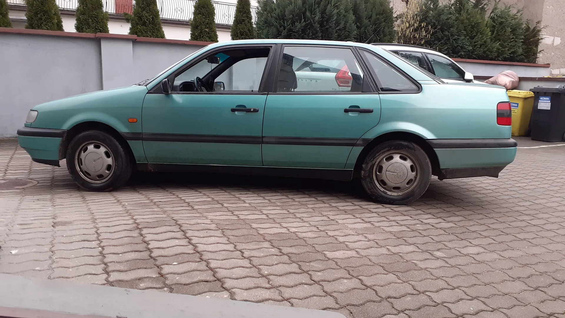 Zdjęcie przedstawia boczny widok jasnoniebieskiego samochodu osobowego typu sedan zaparkowanego na brukowanej powierzchni. Samochód ma czarne listwy nadkoli i srebrne felgi. W tle widoczna jest szara ściana z brązowym wykończeniem, zielone krzewy, częściowo widoczny inny samochód oraz żółty i czarny pojemnik na śmieci z napisem KOMA.