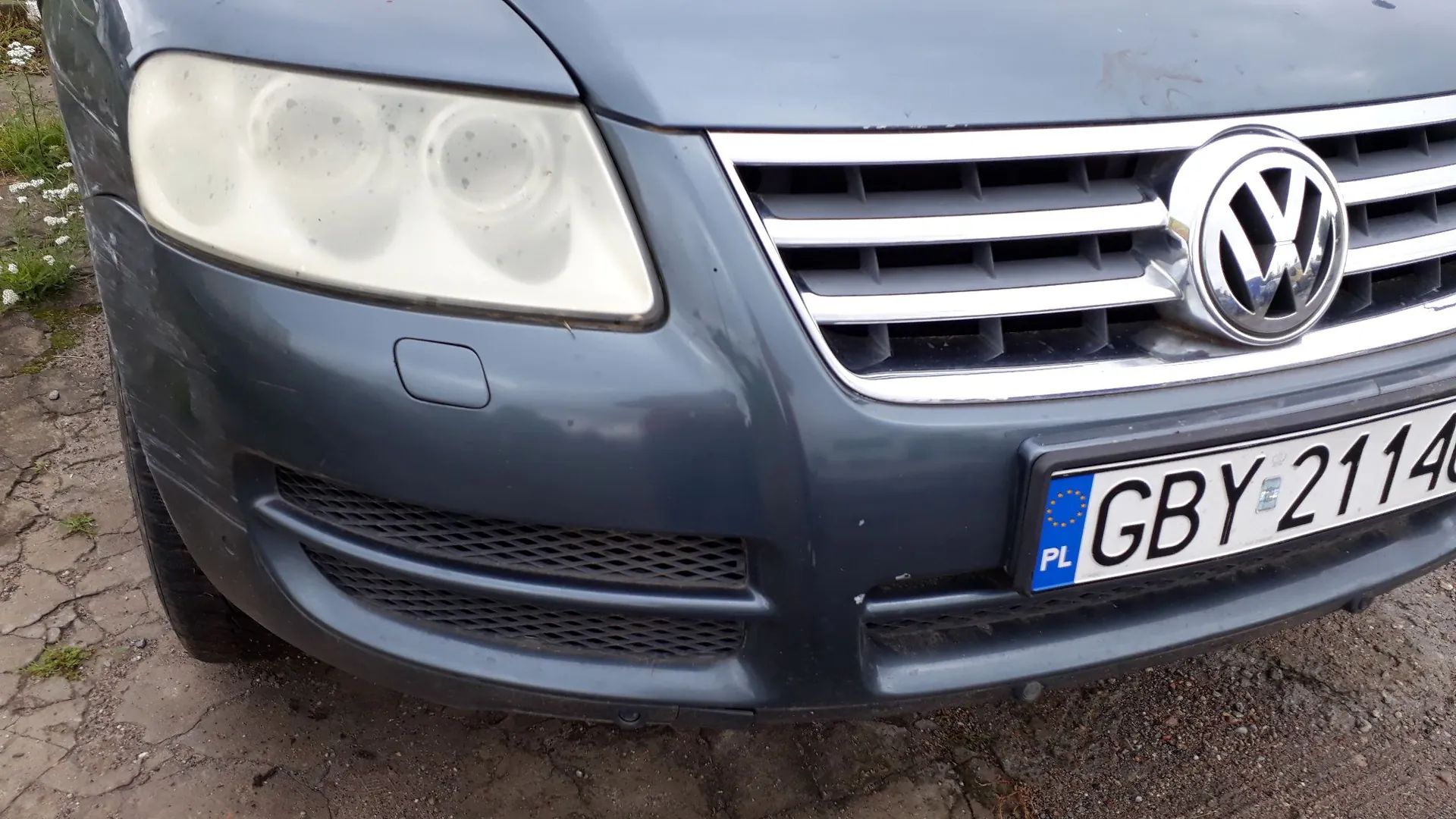 Zbliżenie na przód ciemnoszarego samochodu marki Volkswagen. Widoczny jest lewy reflektor, chromowany grill z logo VW oraz fragment tablicy rejestracyjnej o treści GBY 2114 z oznaczeniem PL.