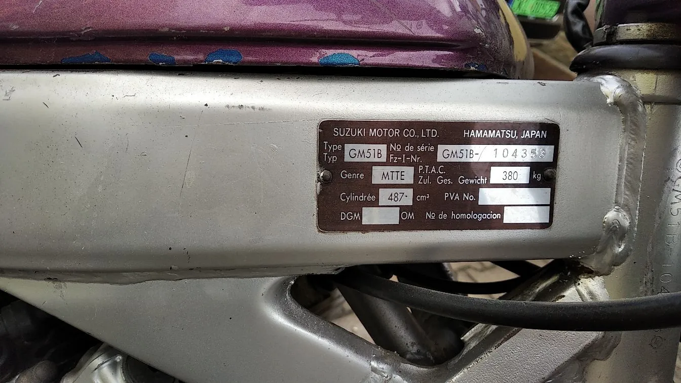 Zbliżenie na srebrną ramę motocykla z przytwierdzoną metalową tabliczką znamionową. Na tabliczce widnieje tekst: 'SUZUKI MOTOR CO., LTD. HAMAMATSU, JAPAN', 'Type Typ GM51B', 'No de série Fz-I-Nr. GM51B- 104353', 'Genre MTTE', 'P.T.A.C. Zul. Ges. Gewicht 380 kg', 'Cylindrée 487 cm³', 'PVA No.', 'DGM', 'OM No de homologacion'. Nad ramą widoczny jest fragment fioletowego lakieru z niebieskimi zdobieniami.