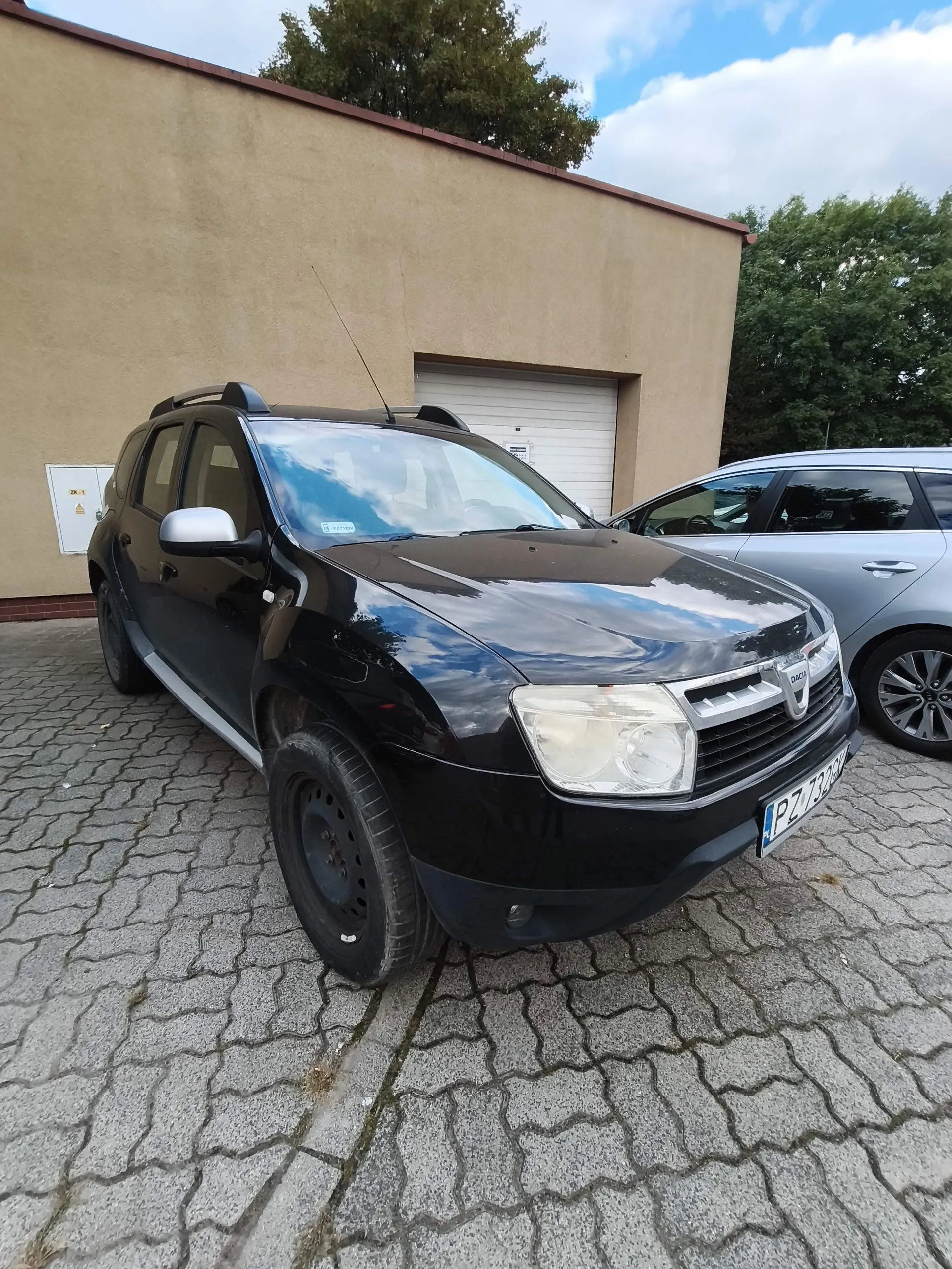 Czarny samochód marki Dacia zaparkowany na kostce brukowej przed beżowym budynkiem z zamkniętą białą bramą garażową. Samochód jest widoczny z przodu i z boku, ma srebrne lusterka boczne i tablicę rejestracyjną o numerze PZ 7322. Obok stoi fragment innego, srebrnego samochodu.