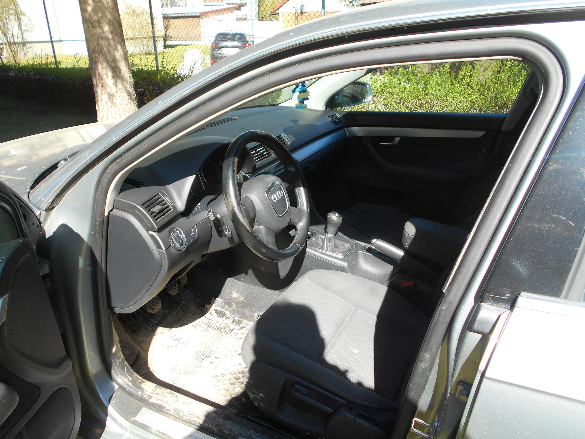 Wnętrze samochodu marki Audi z otwartą drzwiami kierowcy. Widoczne są czarne siedzenia, kierownica z logo Audi, deska rozdzielcza, dźwignia zmiany biegów oraz brudna podłoga z matą. Na zewnątrz samochodu widać drzewo, siatkę ogrodzeniową, zielone krzewy oraz inny samochód w tle. Wewnątrz na desce rozdzielczej znajduje się niebieski przedmiot ozdobny.
