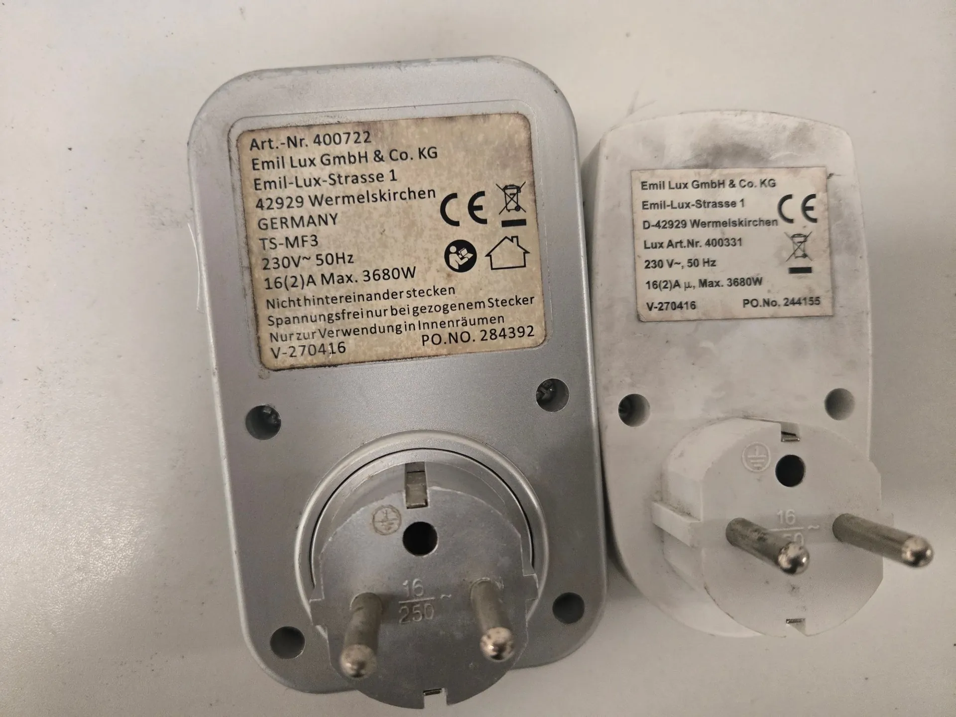 Dwa białe zasilacze elektryczne leżące obok siebie na jasnym tle. Lewy zasilacz ma naklejkę z tekstem: 'Art.-Nr. 400722 Emil Lux GmbH & Co. KG Emil-Lux-Strasse 1 42929 Wermelskirchen GERMANY TS-MF3 230V~ 50Hz 16(2)A Max. 3680W Nicht hintereinander stecken Spannungsfreinur bei beigezogenem Stecker Nur zur Verwendung in Innenräumen V-270416 PO.NO. 284392'. Widoczne są również znaki CE oraz symbol przekreślonego kosza na śmieci. Prawy zasilacz ma mniejszą naklejkę z podobnymi danymi: 'Emil Lux GmbH & Co. KG Emil-Lux-Strasse 1 D-42929 Wermelskirchen Lux Art.Nr. 400331 230 V~, 50 Hz 16(2)A μ, Max. 3680W V-270416 PO.No. 244155' oraz znak CE. Oba urządzenia posiadają wtyczki z dwoma bolcami.