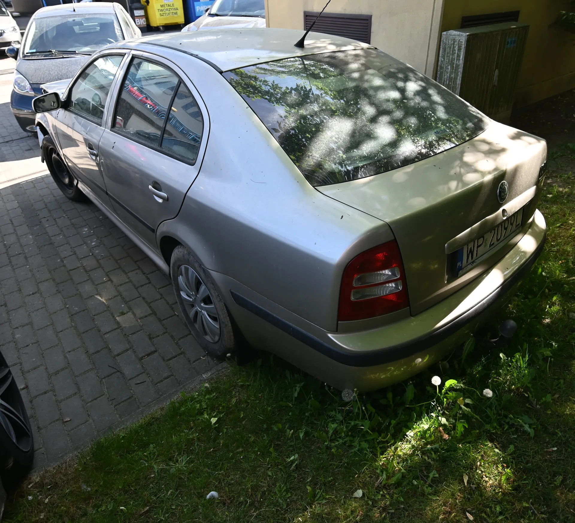 Srebrny samochód osobowy marki Škoda zaparkowany tyłem do obserwatora na brukowanej powierzchni przy zielonej trawie. Widoczna tablica rejestracyjna WP 2099L, czerwone światła tylnego oraz logo marki na bagażniku. W tle inne pojazdy, żółty pojemnik na odpady z napisem METAL ELEKTRO ZŁOM SZCZECIN i BIO oraz fragment budynku z żółtą ścianą i metalową szafą. Na trawie rosną mniszki.