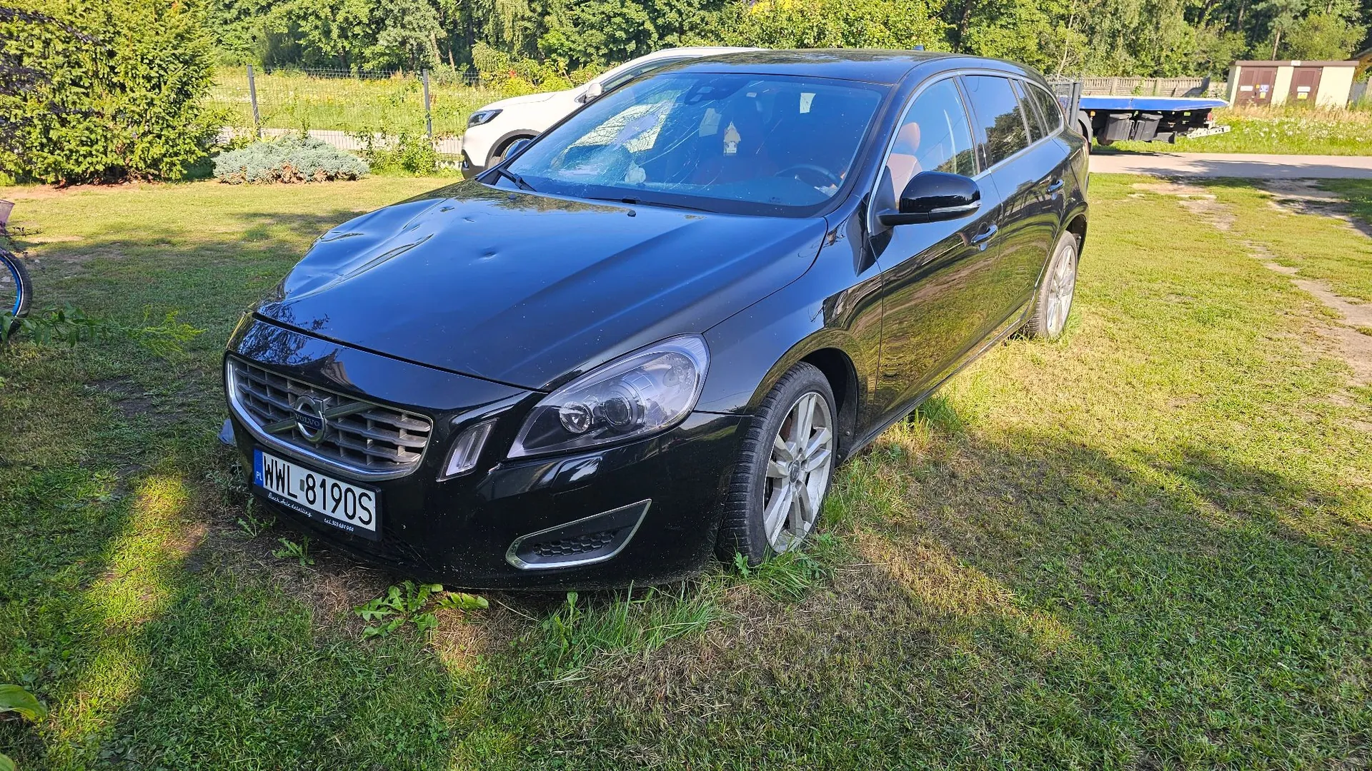 Czarny samochód marki Volvo zaparkowany na trawie. Pojazd ma widoczne uszkodzenia w postaci wgnieceń na masce. Samochód posiada polską tablicę rejestracyjną o numerze WWL 8190S. W tle widoczny jest inny, biały samochód, zielone drzewa oraz fragment niebieskiej przyczepy.