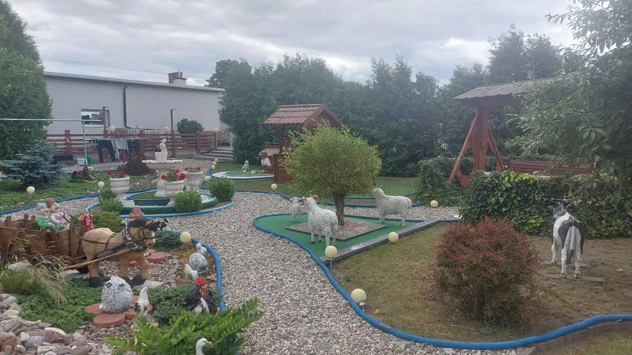 Ogrodowa scena z dekoracyjnymi elementami: posągi owiec, krowy, konia z zaprzęgiem, fontanna z czerwonymi kwiatami, drewniany pawilon z czerwonym dachem, drzewa, krzewy, niebieskie rury wyznaczające ścieżki, kamienista nawierzchnia, budynek z białą elewacją i wiszącym praniem na tle. Niebo jest zachmurzone.