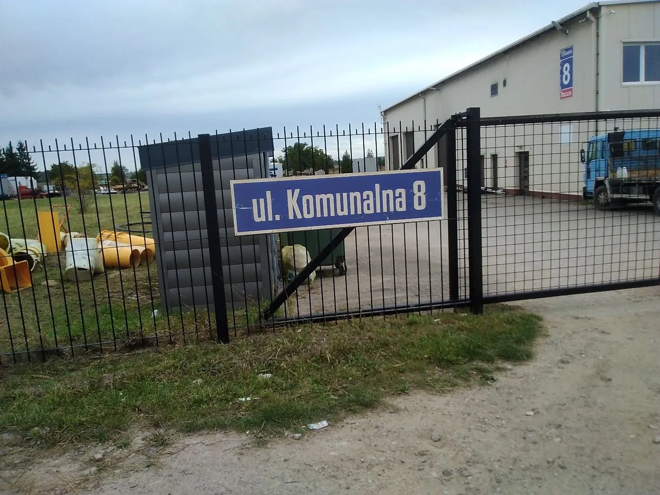 Zdjęcie przedstawia teren przemysłowy z czarnym metalowym ogrodzeniem. Na ogrodzeniu zamocowana jest niebieska tabliczka z białym napisem „ul. Komunalna 8”. Za ogrodzeniem widoczne są żółte rury betonowe, szary modułowy budynek oraz jasny budynek magazynowy w tle. Po prawej stronie stoi niebieska ciężarówka. Podłoże to mieszanka trawy i piaszczystej drogi.