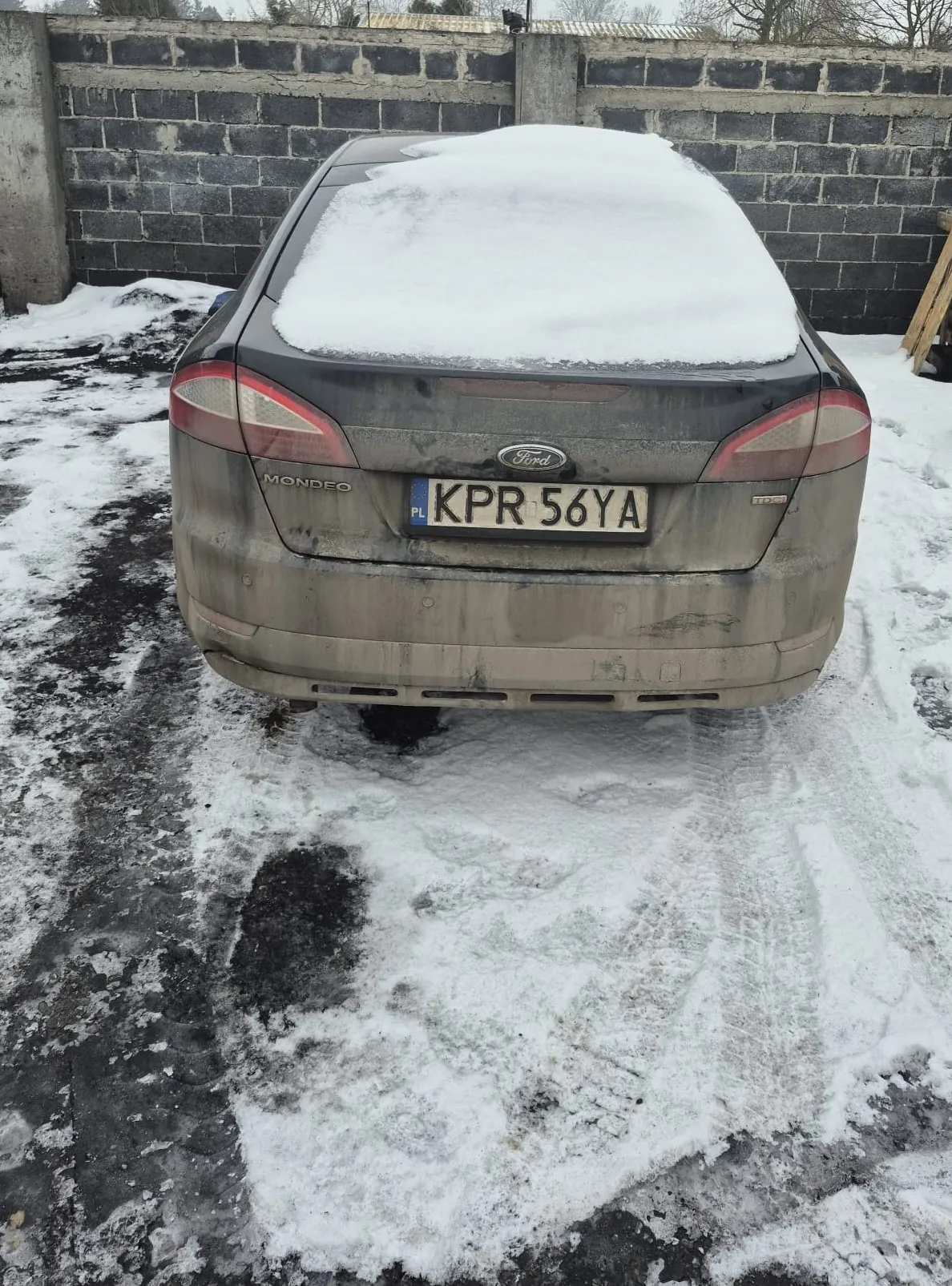 Czarny Ford Mondeo z tablicą rejestracyjną KPR 56YA (PL) stoi na śnieżnej powierzchni z widocznymi odciskami opon. Dach samochodu pokryty jest śniegiem, a nadwozie zabrudzone. W tle betonowy mur z ciemnych bloczków. Na bagażniku widoczne napisy „Mondeo” i logo Ford.