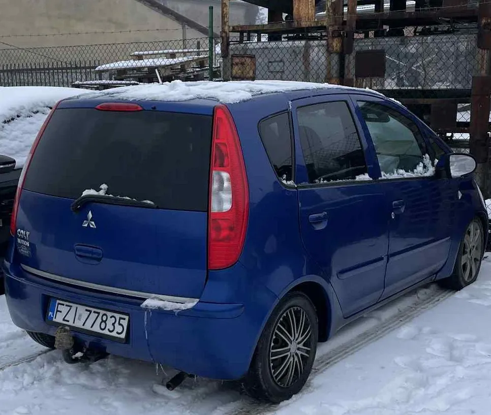 Niebieski samochód Mitsubishi Colt z widocznym napisem 'COLT' na tylnej klapie, stojący na śniegu. Widoczna tablica rejestracyjna PL FZI 77835, czerwone światła tylnego, śnieg na dachu i szybach. W tle siatka ogrodzeniowa, przemysłowe konstrukcje metalowe oraz budynki. Samochód ma czarne felgi i holownik na tylnej osi.