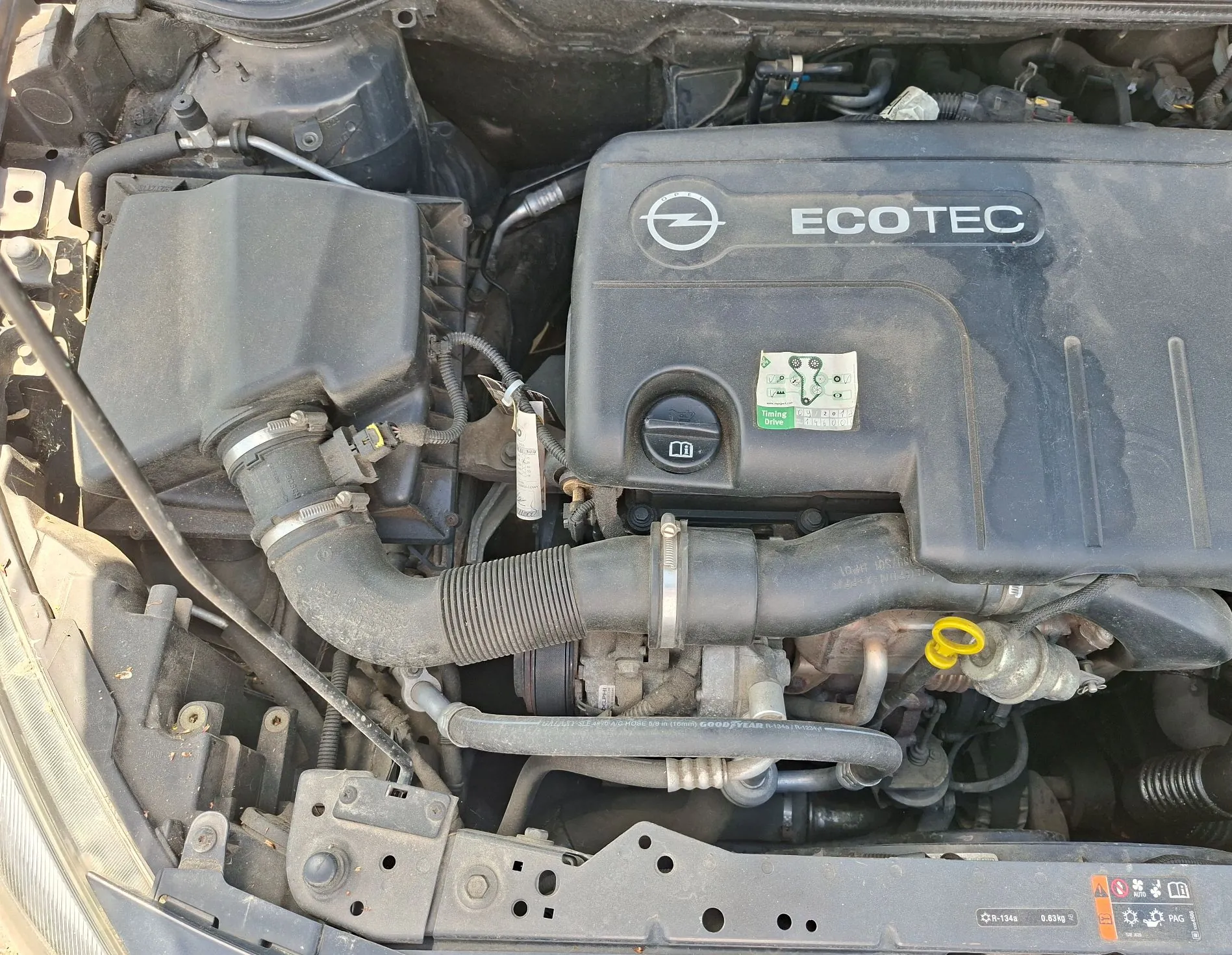 Widok z góry na komorę silnika samochodu. Centralnie znajduje się czarna plastikowa osłona silnika z logo marki Opel oraz białym napisem 'ECOTEC'. Po lewej stronie widoczna jest obudowa filtra powietrza połączona z silnikiem grubym, gumowym wężem. W dolnej części zdjęcia widoczne są elementy konstrukcyjne nadwozia, przewody oraz żółty uchwyt bagnetu do pomiaru poziomu oleju. Na osłonie silnika naklejona jest mała, zielono-biała etykieta informacyjna.