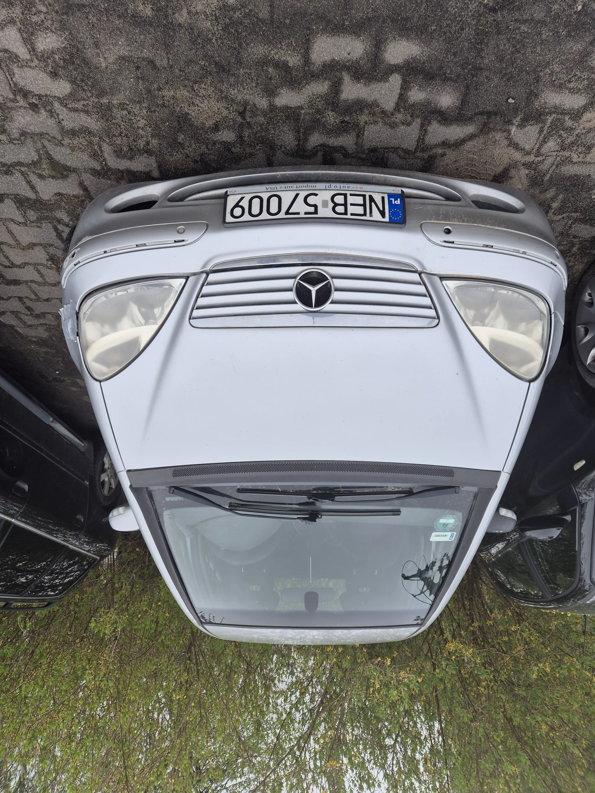 Srebrny samochód marki Mercedes-Benz z tablicą rejestracyjną NEB 57009. W tle czarny samochód, zielone drzewa i brukowana powierzchnia. Tablica rejestracyjna zawiera dodatkowo oznaczenie PL oraz napisy po prawej stronie.