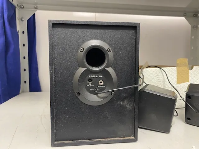 Czarny głośnik z przodu, z okrągłym portem basowym i napisem 'SBS 2.1 300' oraz oznaczeniami 'LINE IN' i 'SUBWOOFER' pod portem. Po prawej stronie znajdują się dwa mniejsze czarne głośniki połączone przewodami. Tło: biała powierzchnia, niebieska zasłona po lewej, metalowe półki i biała ściana z taśmą klejącą.