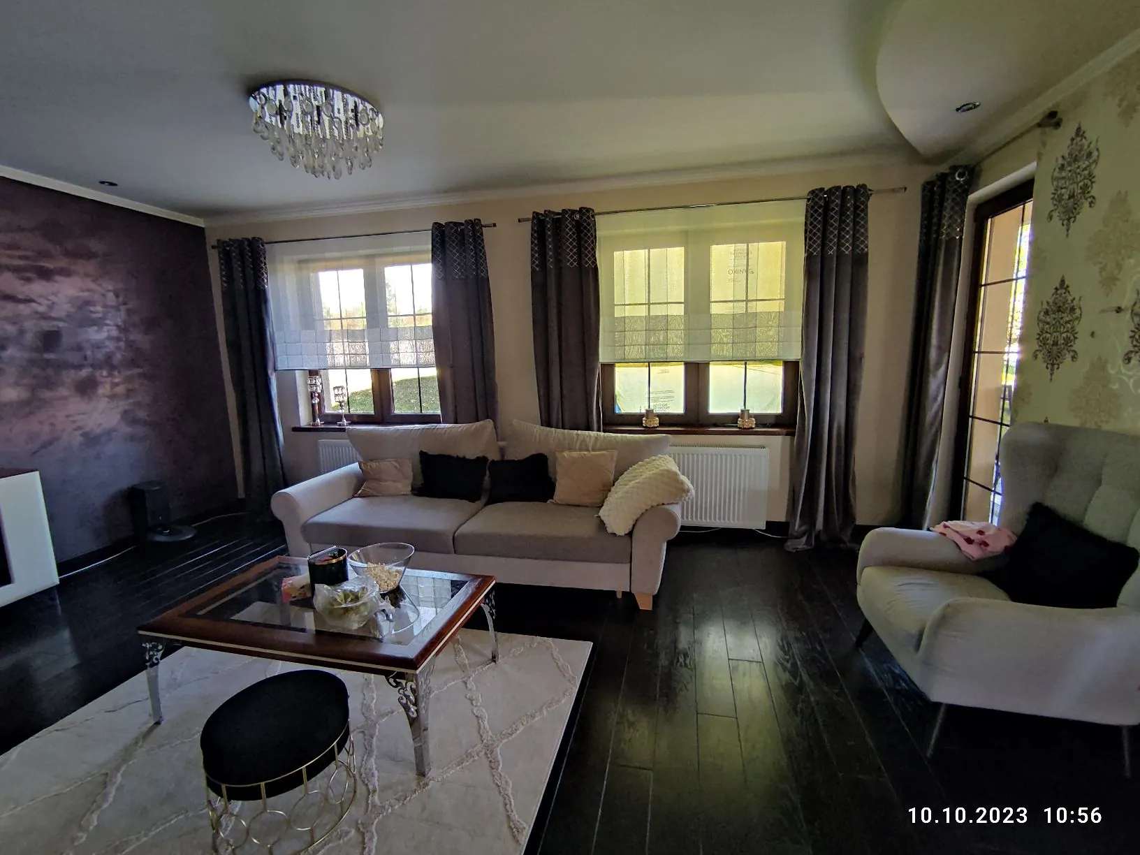 Wnętrze salonu z ciemną drewnianą podłogą. W centrum znajduje się jasnoszara sofa z kilkoma poduszkami w kolorach czarnym i beżowym oraz białym kocem. Przed sofą stoi szklany stolik kawowy na ozdobnych srebrnych nogach, na którym znajdują się dekoracje i miseczka. Pod stolikiem leży biały dywan, a obok niego czarny puf z złotym stelażem. Po prawej stronie stoi jasnoszary fotel z czarną poduszką. W tle widoczne są dwa okna z brązowymi ramami, zasłonięte szarymi zasłonami i rzymskimi roletami. Po lewej stronie znajduje się ściana w ciemnym, fioletowo-brązowym kolorze. Na suficie wisi kryształowy żyrandol. W prawym dolnym rogu widnieje data i godzina: 10.10.2023 10:56.