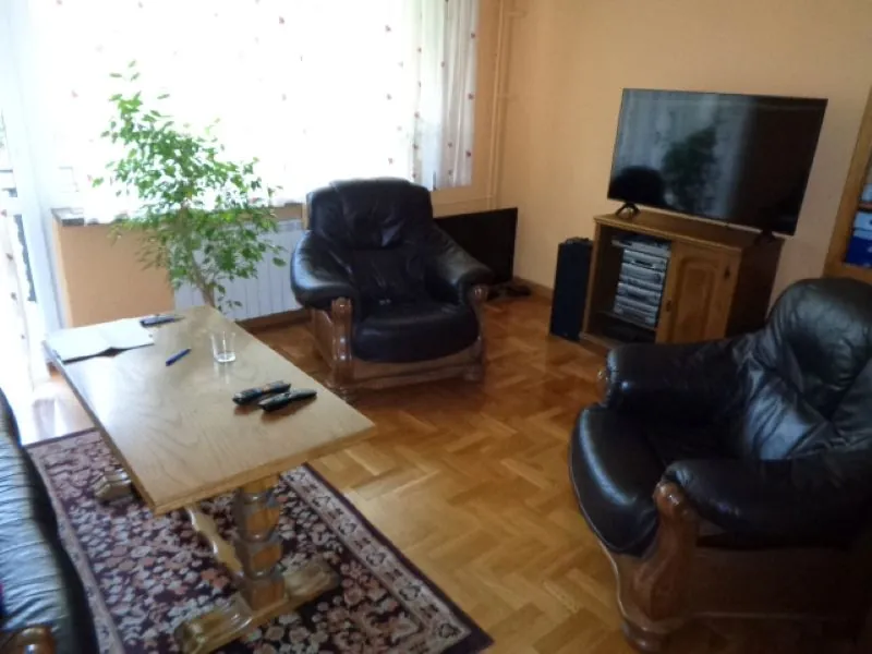 Wnętrze pokoju z drewnianą podłogą ułożoną w jodełkę. W pomieszczeniu znajdują się dwa duże, czarne skórzane fotele z drewnianymi podłokietnikami. Po lewej stronie stoi jasny, drewniany stół, na którym leży otwarta książka, długopis i dwa piloty. Pod stołem rozłożony jest wzorzysty dywan w odcieniach czerwieni i granatu. W rogu pokoju znajduje się zielona roślina doniczkowa. Po prawej stronie stoi drewniana szafka RTV z telewizorem oraz sprzętem audio. W tle widoczne jest okno z białymi firankami.