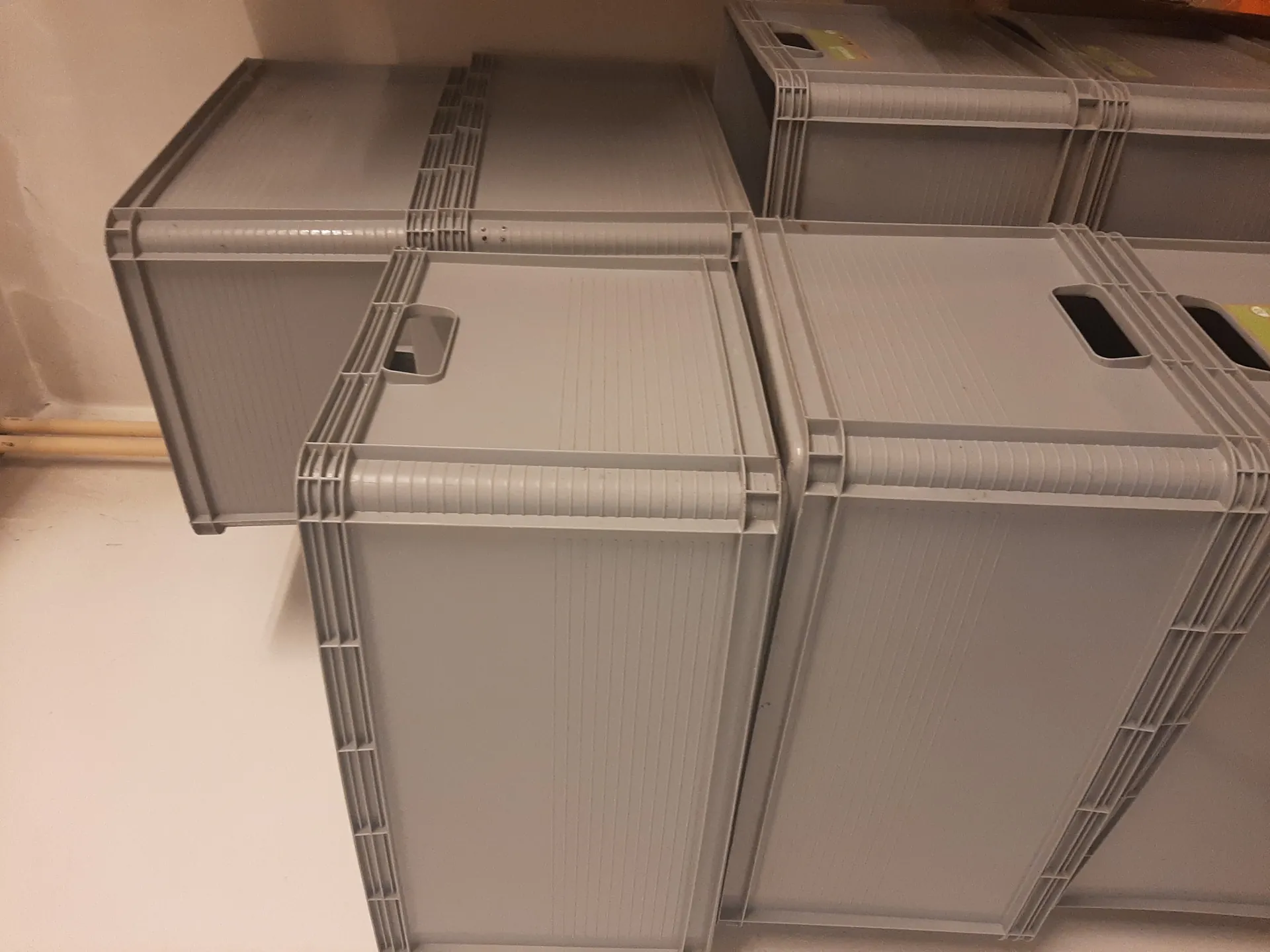 Stos szarych plastikowych pojemników z uchwytami, ułożonych pionowo na sobie. Pojemniki mają prostokątny kształt, gładkie ściany z poziomymi liniami i otwory uchwytowe. W tle biała ściana z widoczną pionową rurą. Niektóre pojemniki mają częściowo widoczne etykiety z kolorowymi elementami.