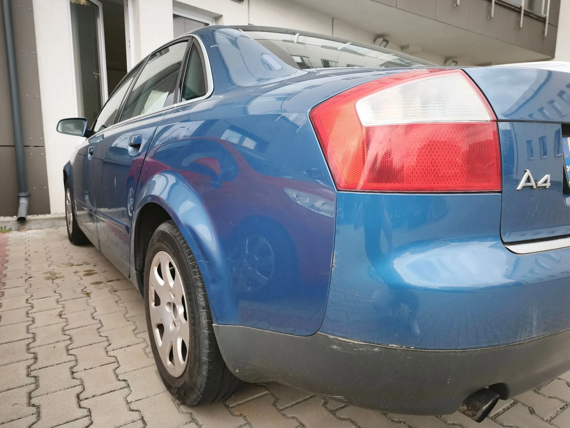 Niebieski samochód osobowy marki Audi A4 widziany od tyłu z boku. Na klapie bagażnika widoczny jest srebrny napis 'A4'. Auto zaparkowane jest na kostce brukowej obok jasnego budynku.
