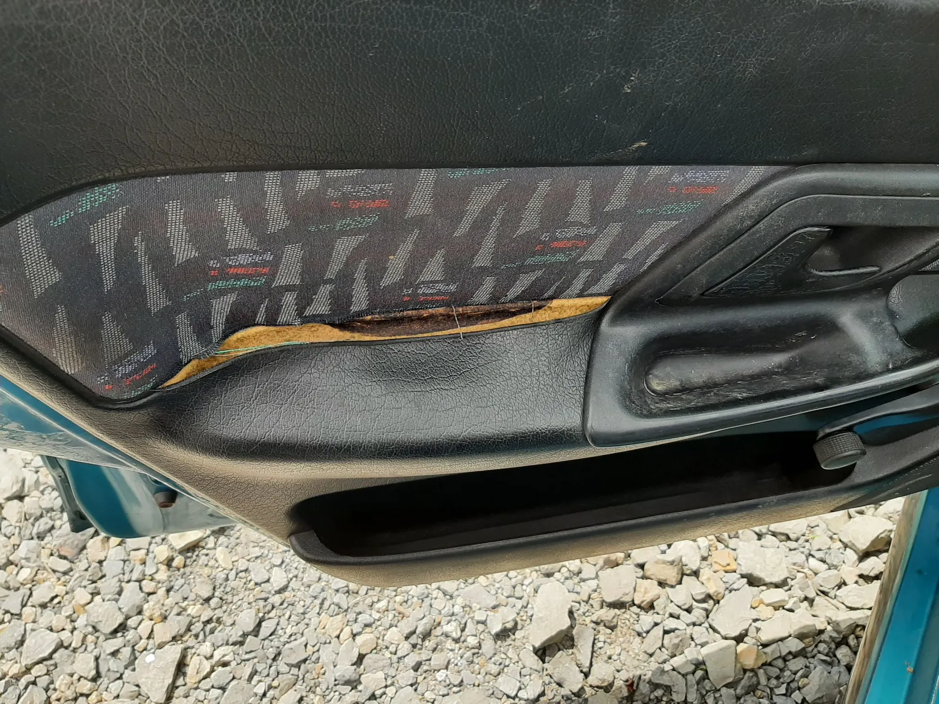 Zbliżenie na wewnętrzną część drzwi samochodu. Widoczny jest czarny plastikowy panel z kieszenią na dole oraz fragment tapicerki materiałowej w kolorze ciemnoszarym z geometrycznym wzorem. W materiale znajduje się poziome rozdarcie, przez które prześwituje żółta gąbka wypełniająca. Obraz jest zorientowany poziomo.