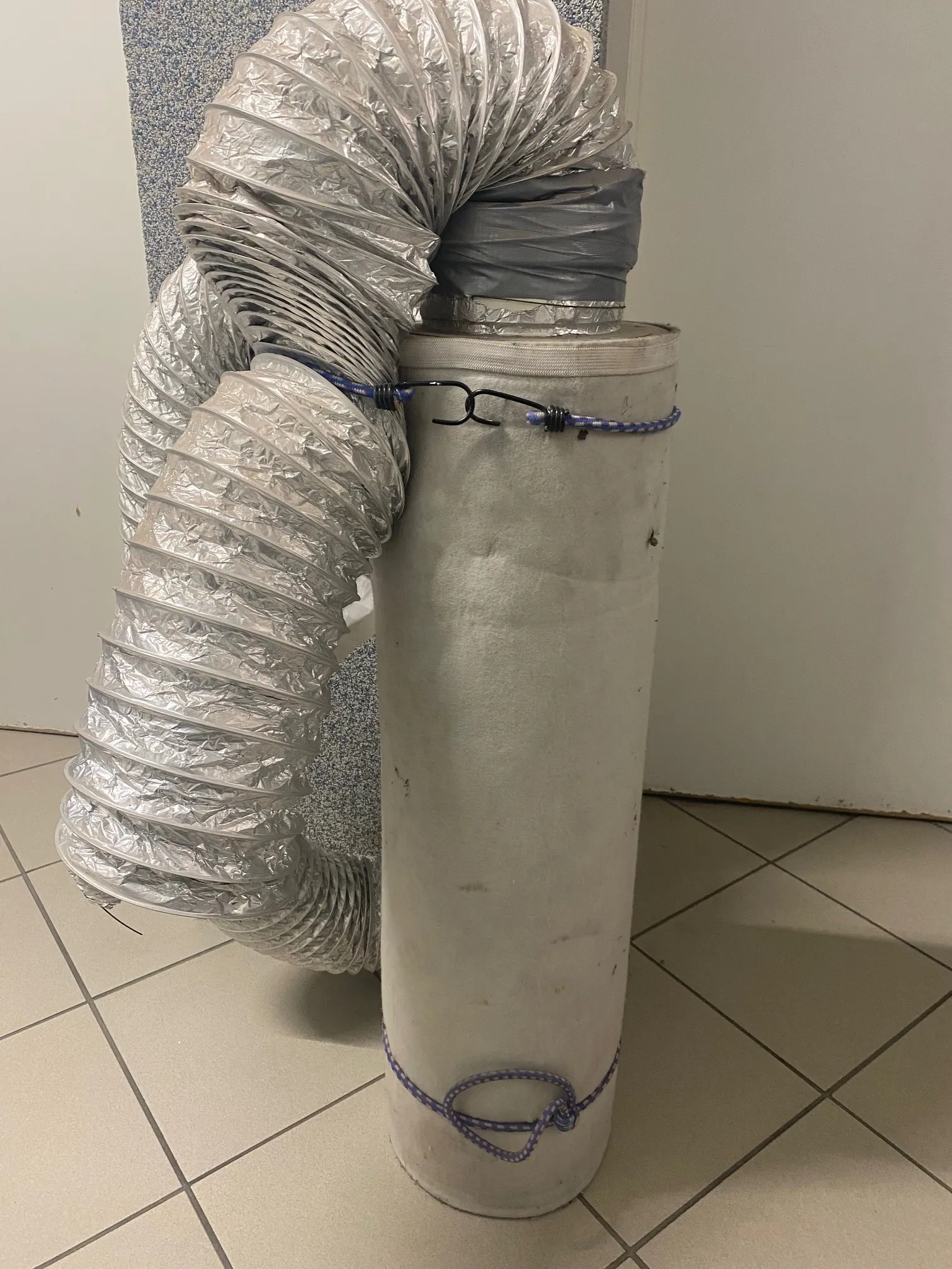 Wysoki, biały lub szary cylinder z materiału przypominającego płótno, przytwierdzony niebiesko-białą sznurówką w dwóch miejscach. Na górze cylindra znajduje się srebrny, elastyczny kanał wentylacyjny z metalizowaną powłoką, częściowo owinięty szarą taśmą. Tło stanowi podłoga z jasnych płytek ceramicznych i biała ściana z fragmentem tekstury przypominającej panel. Brak czytelnego tekstu.