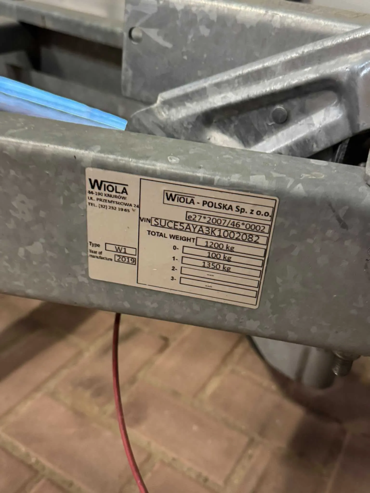 Zbliżenie na metalową tabliczkę znamionową przymocowaną do srebrnej, ocynkowanej konstrukcji. Na tabliczce znajduje się tekst: 'WiOLA 84-190 KNURÓW UL. PRZEMYSŁOWA 24 TEL. (32) 232 1965', 'WiOLA - POLSKA Sp. z o.o.', 'e27*2007/46*0002', 'VIN SUCES5AYA3K1002082', 'TOTAL WEIGHT 1200 kg'. Poniżej znajduje się tabela z polami: 'Type W1', 'Year of manufacture 2019' oraz wiersze z wagami: '0- 100 kg', '1- 1350 kg'.