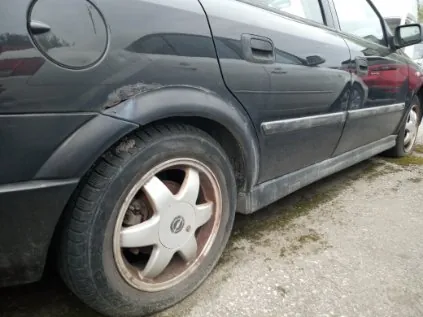 Czarny samochód widziany z boku, z naciskiem na tylne koło i zarysowaną błotnik. Koło ma srebrną felgę z logo w środku. Na błotniku widoczne są uszkodzenia i zabrudzenia. Tło stanowi nierówna, zagruntowana powierzchnia z fragmentem czerwonego samochodu w tle.