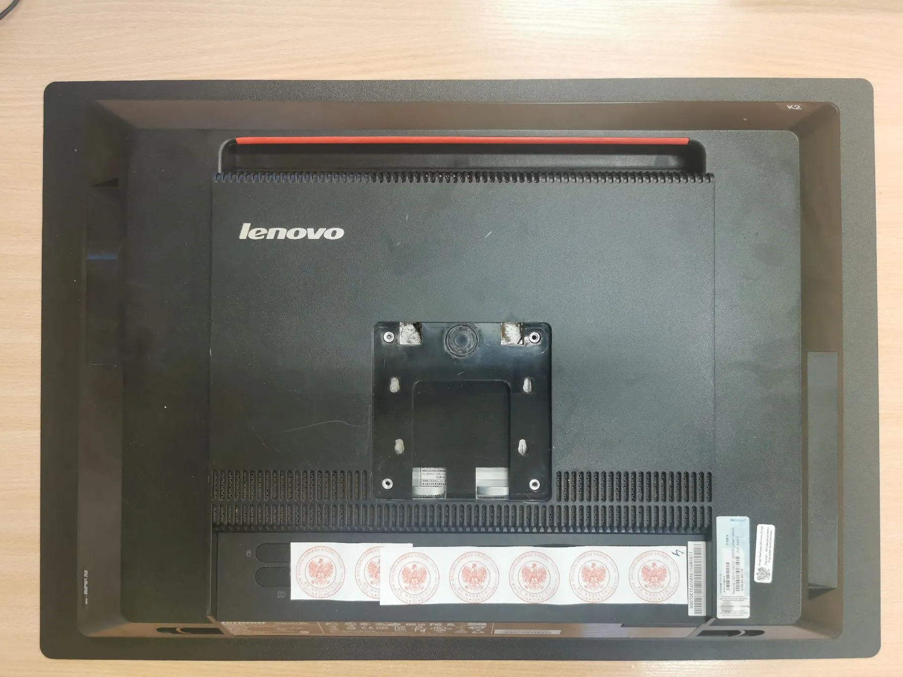 Tylna obudowa czarnego monitora marki Lenovo. Na środku znajduje się biały napis 'lenovo'. Poniżej widoczny jest kwadratowy uchwyt VESA do montażu na ścianie. W dolnej części obudowy znajdują się kratki wentylacyjne oraz biała naklejka z kodami kreskowymi i informacjami technicznymi. Na dole obudowy widnieje rząd okrągłych, czerwono-białych pieczęci urzędowych z godłem Polski.