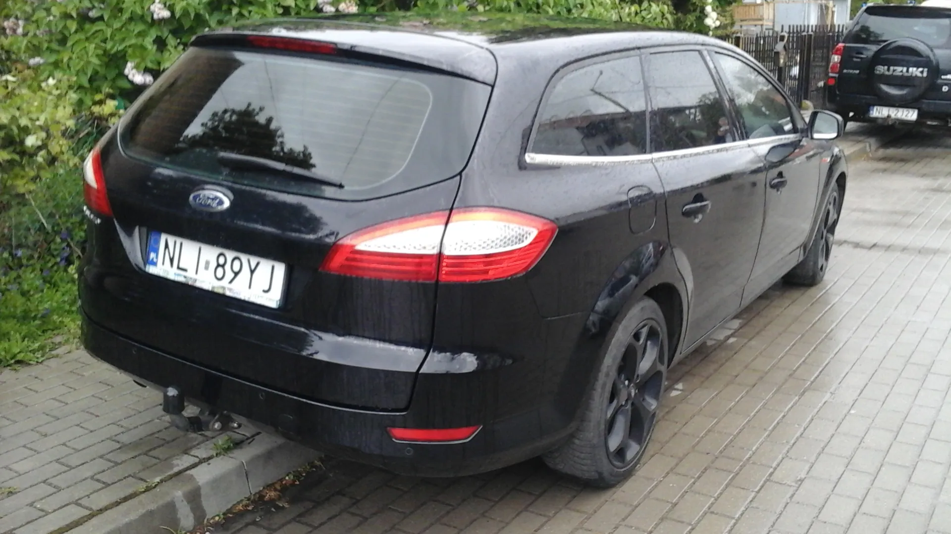 Czarny samochód osobowy marki Ford, widok z tyłu, zaparkowany na brukowanej powierzchni. Widoczny tabliczka rejestracyjna PL NL 89YJ, czerwone światła tylnego, logo Ford na bagażniku. W tle zielone krzewy, czarny samochód Suzuki z tabliczką NL 2127 oraz metalowe ogrodzenie. Podłoga z cegieł, fragment chodnika z krawężnikiem.