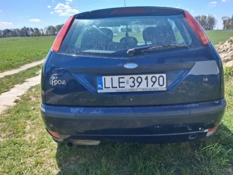 Zdjęcie przedstawia tył ciemnoniebieskiego samochodu marki Ford Focus zaparkowanego na trawie. Widoczna jest tablica rejestracyjna LLE 39190 z polskim flagą i literą PL po lewej stronie. Na lewej stronie bagażnika znajduje się napis focus. W tle rozległe pola, droga gruntowa oraz niebo z kilkoma chmurami. Samochód stoi na trawie, a w oddali widoczne są drzewa.