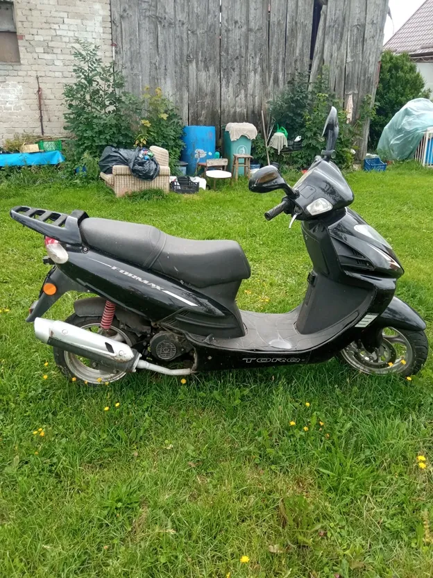 Czarny skuter marki Toro, model Formax, zaparkowany na zielonej trawie. Pojazd jest widoczny z profilu, posiada czarną kanapę, srebrny wydech oraz bagażnik z tyłu. W tle znajduje się drewniany płot, krzewy, niebieska beczka oraz inne przedmioty gospodarcze.