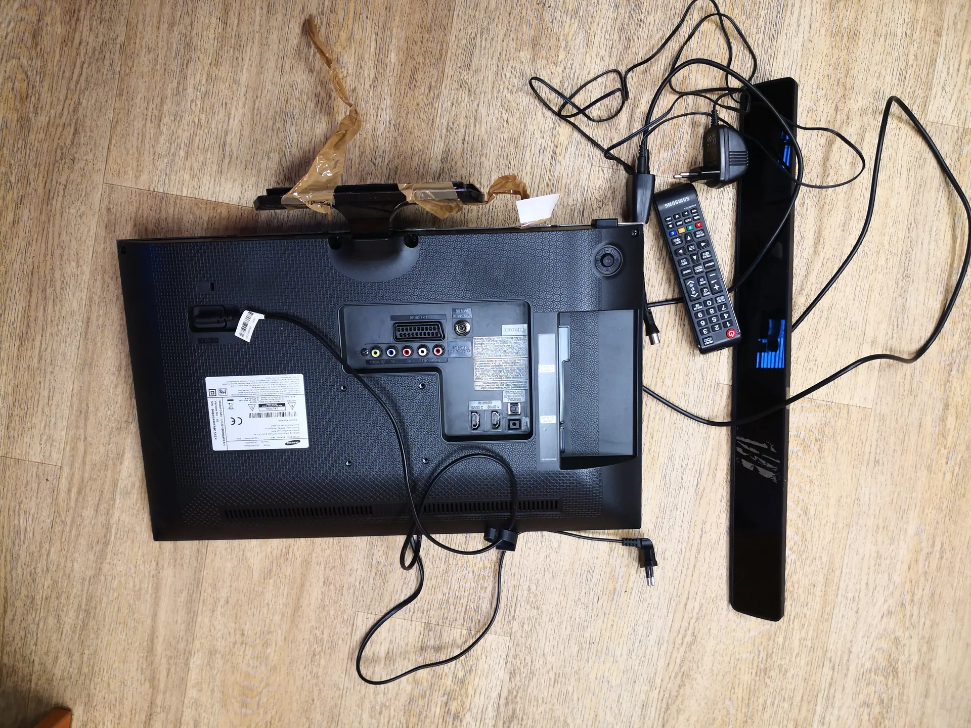 Zdjęcie przedstawia odwróconą tylną stronę czarnego telewizora leżącego na podłodze z drewnianą fakturą. Widoczne są porty wejściowe (w tym HDMI, VGA, gniazda audio), białe etykiety z niewyraźnym tekstem oraz podłączony kabel zasilający. Obok telewizora znajduje się czarny pilot z przyciskami, zasilacz i rozłożone przewody. Na górze telewizora leży brązowa taśma klejąca. Całość ułożona jest na podłodze z widocznym wzorem drewna.