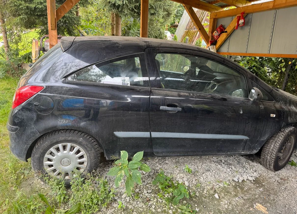 Czarny, zakurzony samochód osobowy typu hatchback zaparkowany pod drewnianym zadaszeniem na żwirowym podłożziu. Pojazd jest widoczny z profilu od strony kierowcy. W tle widać zielone rośliny i fragmenty konstrukcji zadaszenia, na której zawieszona jest czerwona gaśnica.