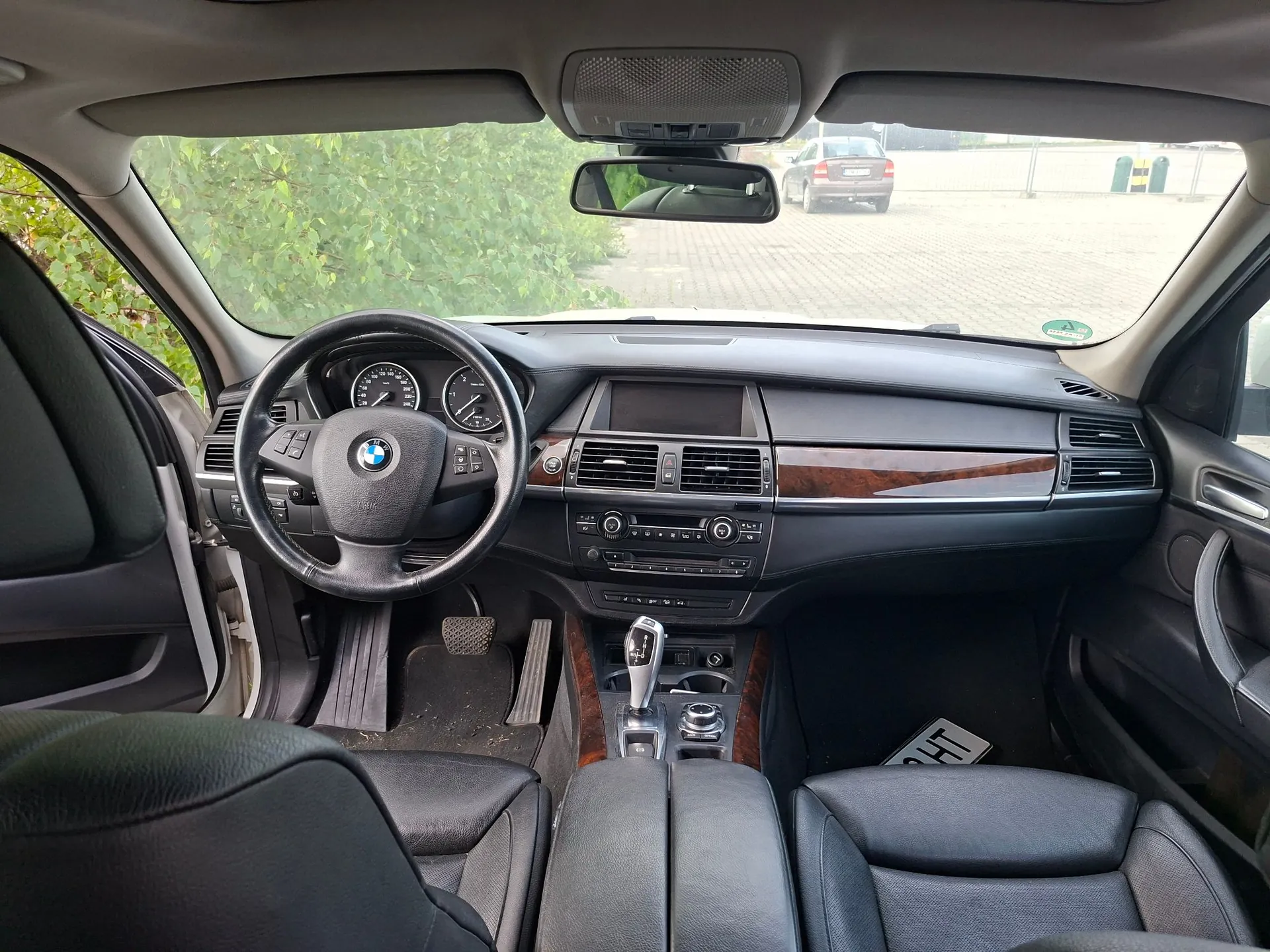 Wnętrze samochodu marki BMW widziane z perspektywy tylnego siedzenia. Widoczna jest czarna skórzana tapicerka, kierownica z logo BMW, deska rozdzielcza z drewnianymi wstawkami, panel centralny z ekranem i przyciskami oraz dźwignia zmiany biegów. Przez przednią szybę widać fragment zielonych krzewów oraz zaparkowany w oddali srebrny samochód.