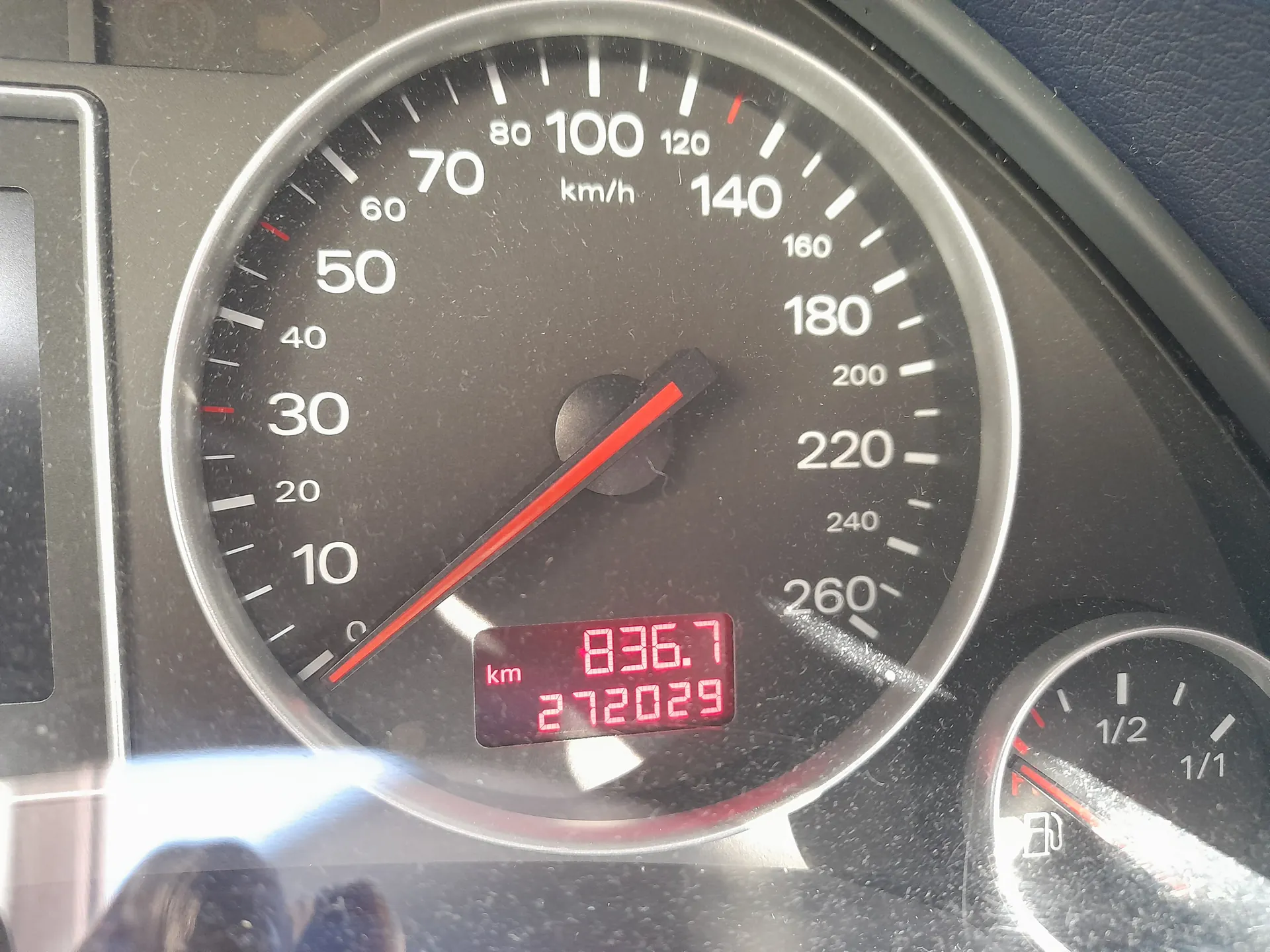 Zbliżenie na zegar samochodowy z prędkościomierzem wskazującym prędkość w km/h. Skala prędkościomierza od 0 do 260 km/h z białymi cyframi i czerwoną strzałką wskazującą około 50 km/h. Poniżej znajduje się czerwony wyświetlacz cyfrowy z napisem "km" oraz wartościami "836.7" i "272029". Po prawej stronie widoczny jest wskaźnik paliwa z oznaczeniami 1/2 i 1/1 oraz symbolem zbiornika paliwa. Tło zegara jest czarne, a obramowanie srebrne.